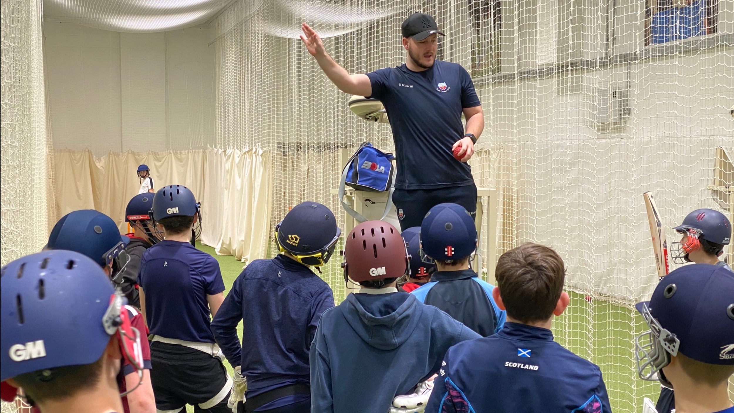 A coach giving instructions to young cricket players with helmets, indoors, during a training session.