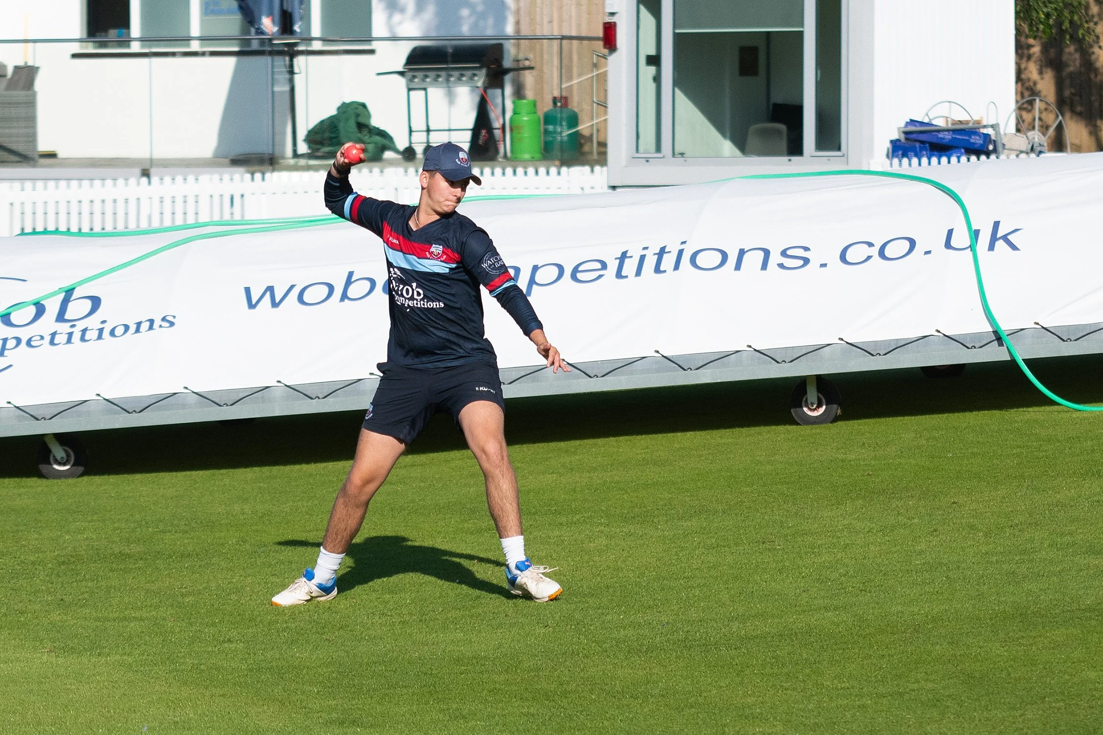 A young male athlete in sportswear and a cap is standing on a grassy field, holding a red object, possibly preparing for a shot-put event, with a large white banner and modern building in the background.