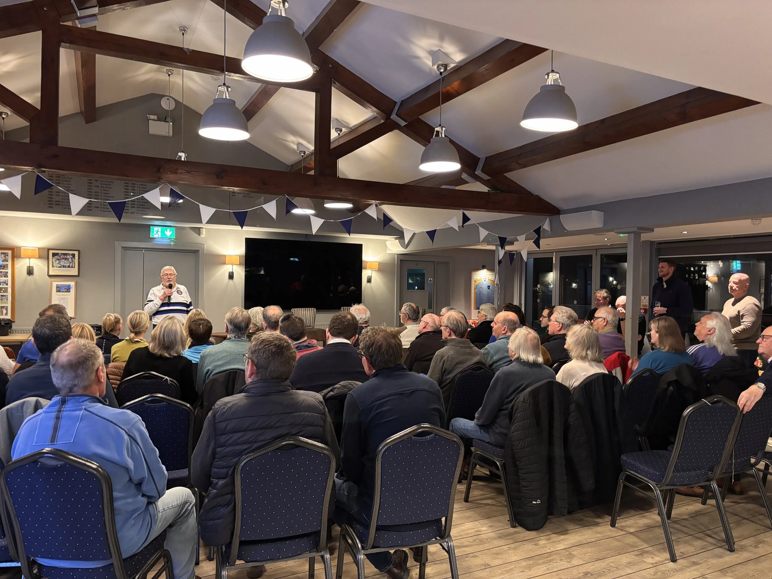 A speaker stands at the front of a room filled with seated audience members. The room has wooden beams on the ceiling, hanging lights, and bunting decorations. The audience is listening attentively to the speaker.