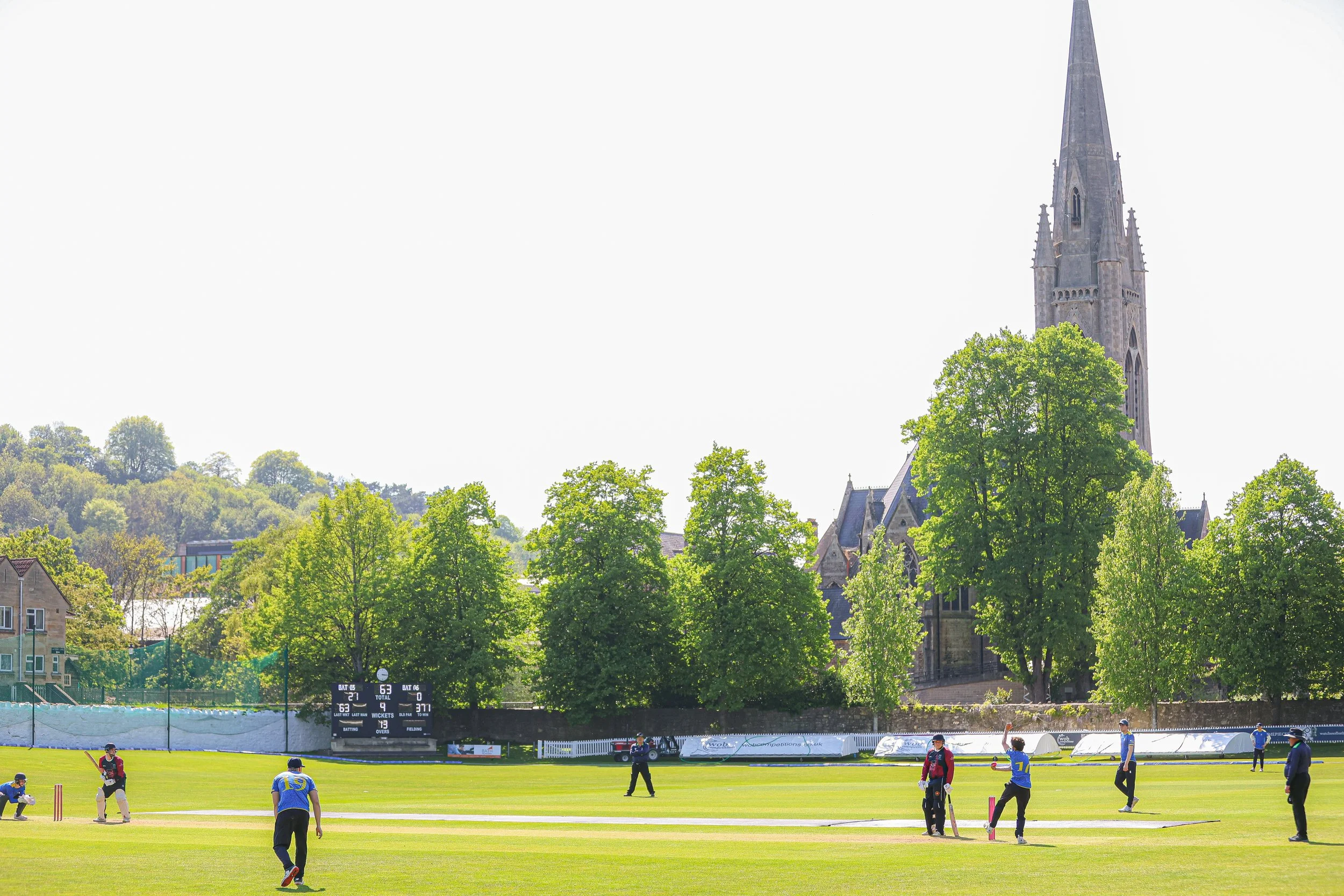 Cricket players on a field with a scoreboard and a church with a tall steeple in the background, surrounded by green trees under a bright sky.