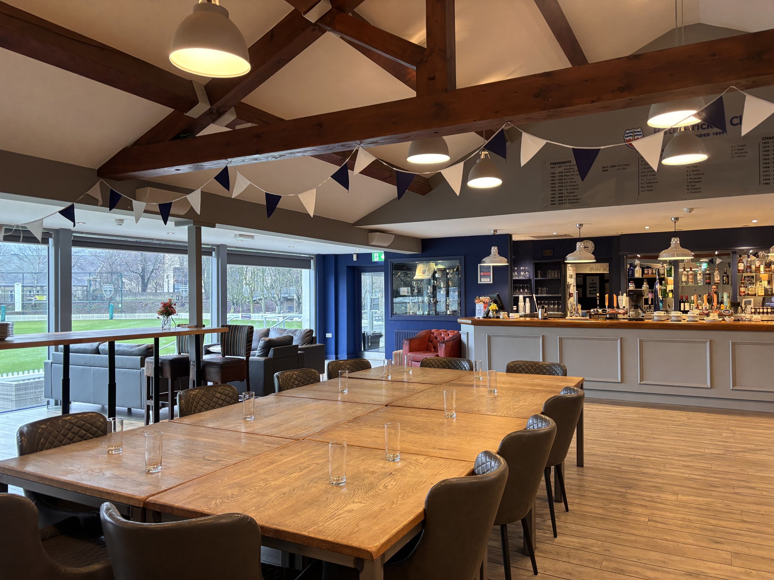 A spacious restaurant or pub interior with wooden beams on the ceiling, large windows, a bar area with various bottles, and seating options including chairs and sofas. There's a decorative garland with navy and white bunting hanging from the ceiling.