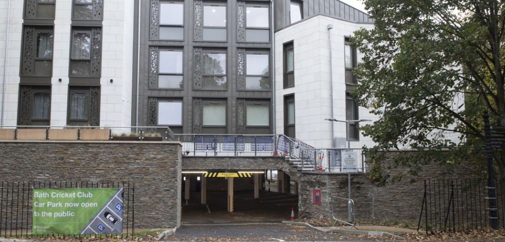 The image shows a multi-story parking garage entrance under an apartment building, with a sign for Bath Cricket Club parking and a tree on the right side.