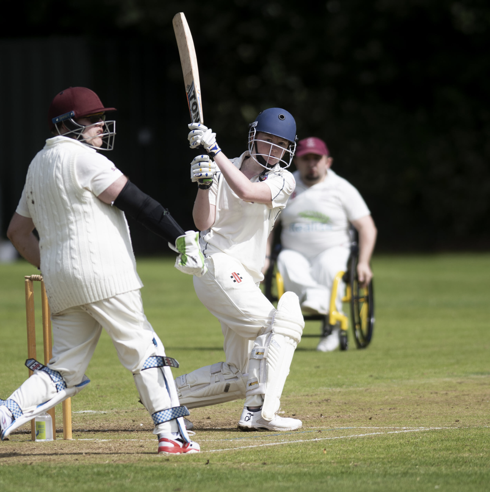 A cricket player in white uniform batting, with a wicketkeeper and a fielder in the background on a grassy field.