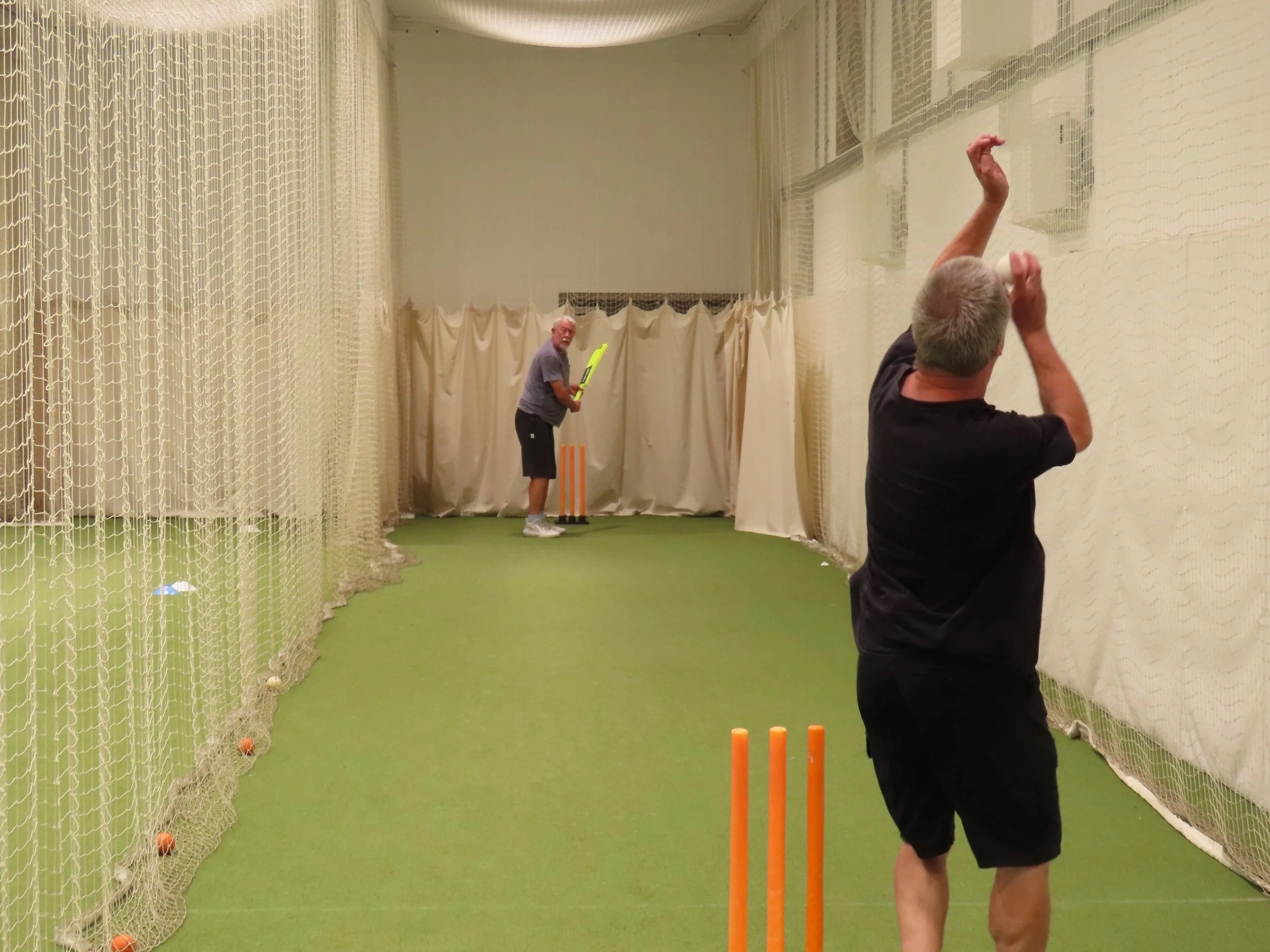 Two men playing cricket indoors in a netted area with white curtains. One man is holding a cricket bat, preparing to hit a ball, while the other man is facing away, with arms raised, possibly bowling or fielding.