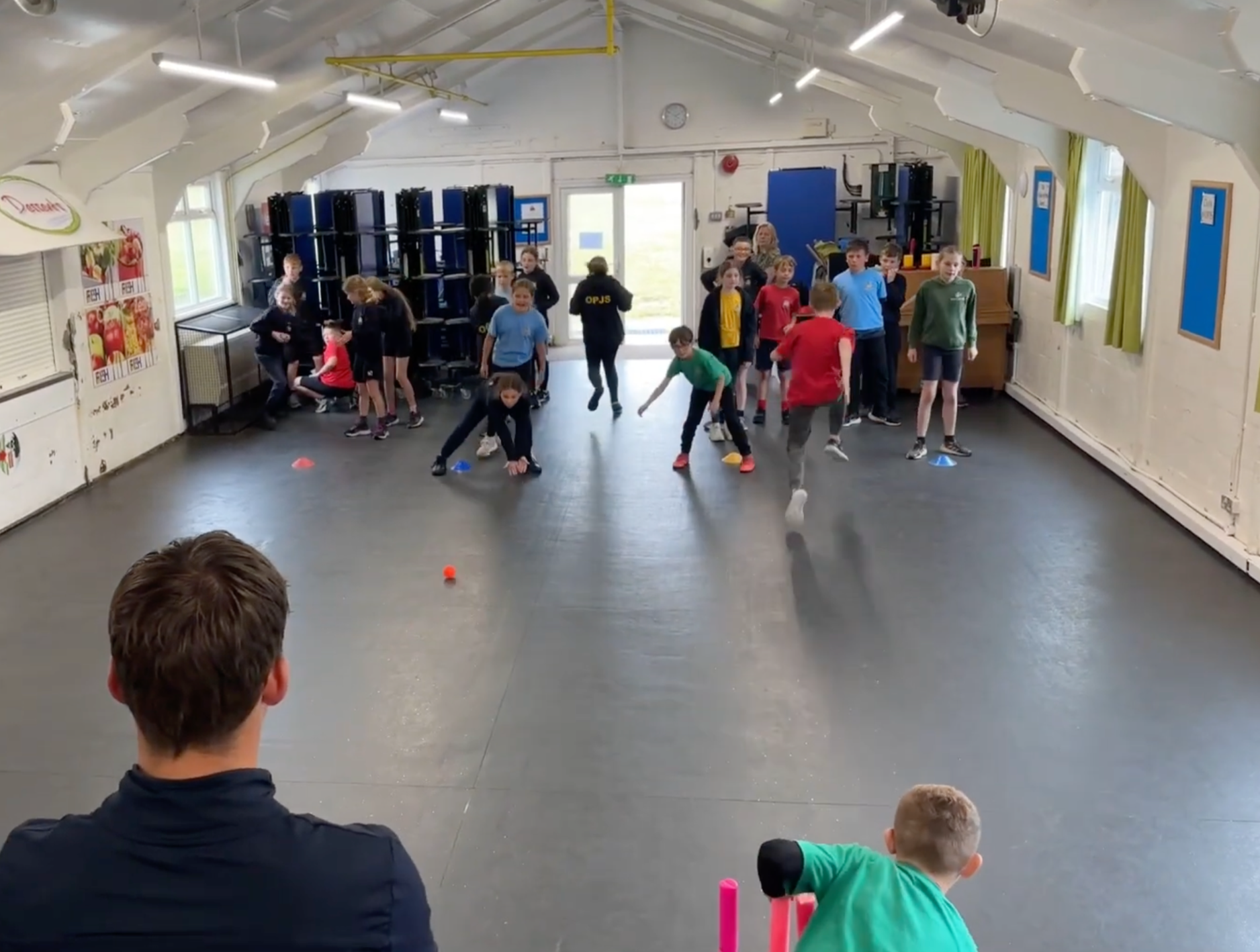 Children participating in an indoor activity, some reaching for objects on the floor, in a room with blue chairs stacked against the wall, windows with green curtains, and adults supervising.