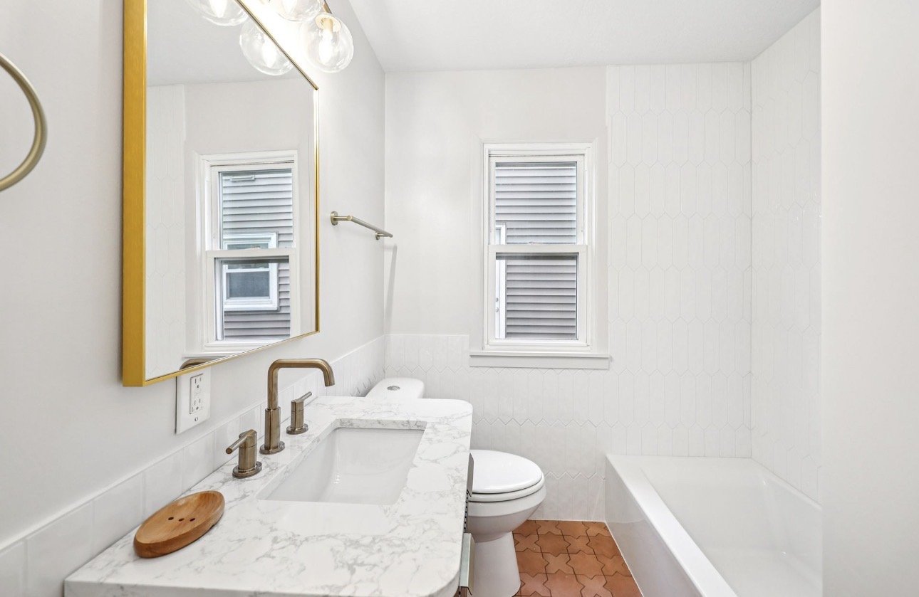 A bright bathroom with a vanity mirror, marble countertop, and a wooden tray. There is a window with blinds, a toilet, and a bathtub. The walls are painted white, and the floor has reddish-brown tiles.