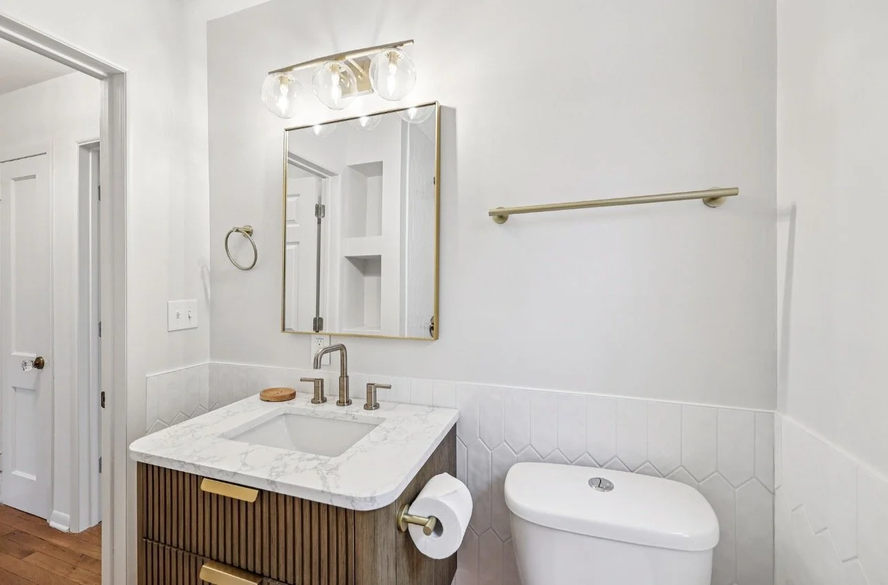 Modern bathroom with white walls, a marble-top vanity with brass handles, a rectangular mirror, wall-mounted lighting fixture, brass towel ring, toilet with a spare toilet paper roll, and a brass towel bar on the wall.