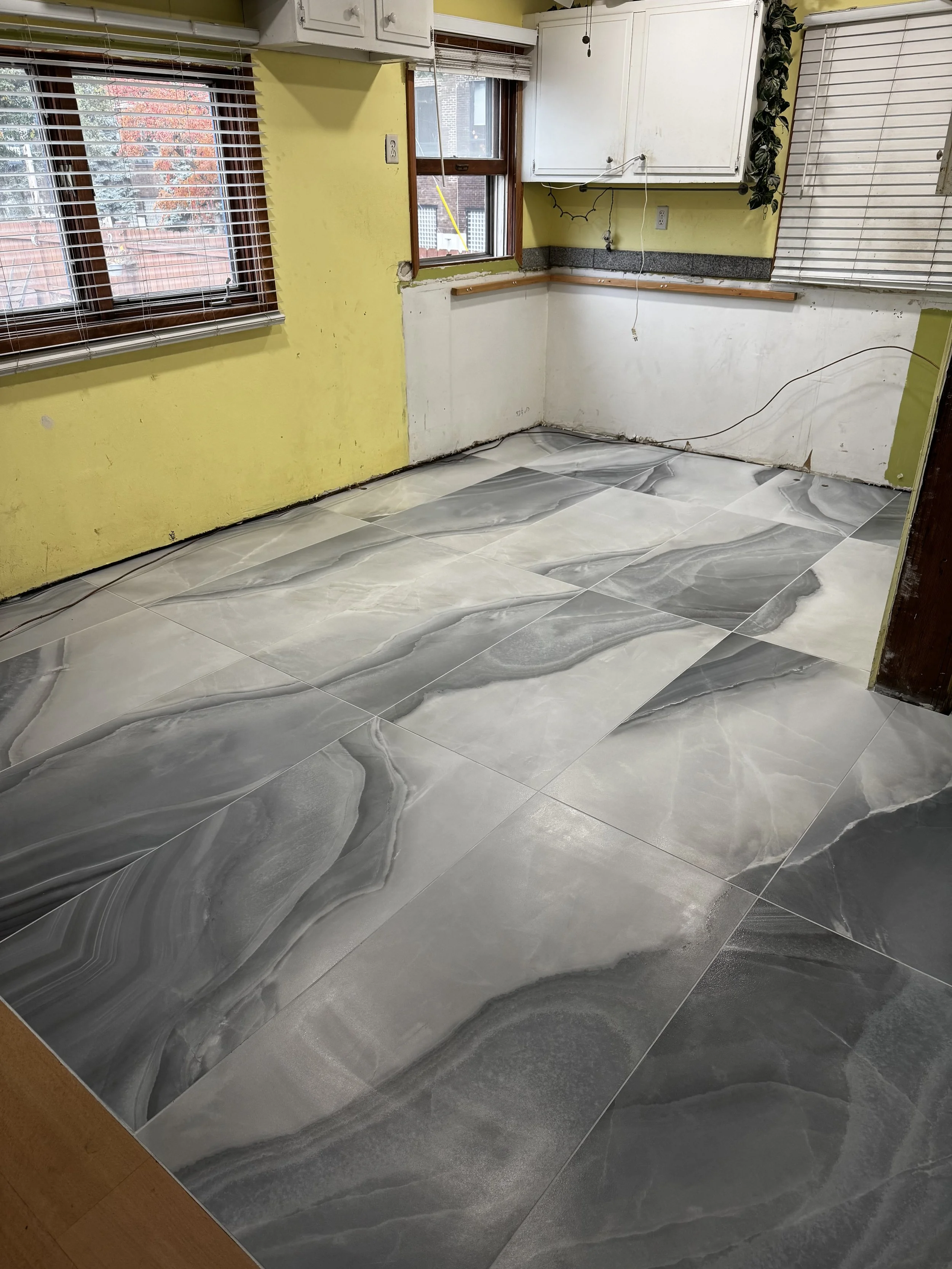 Room with marble-patterned gray and white tiles on the floor, yellow and white walls, two windows with blinds, and white kitchen cabinets.