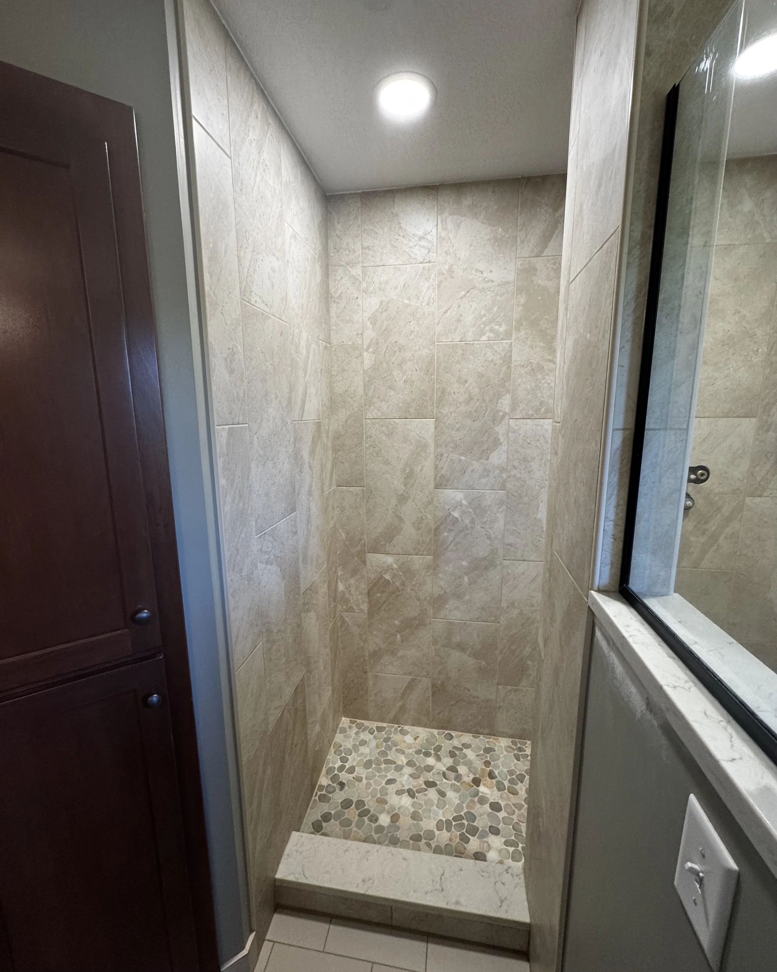 Empty walk-in shower with beige tiled walls and pebble stone floor, between a wooden cabinet and a bathroom mirror.