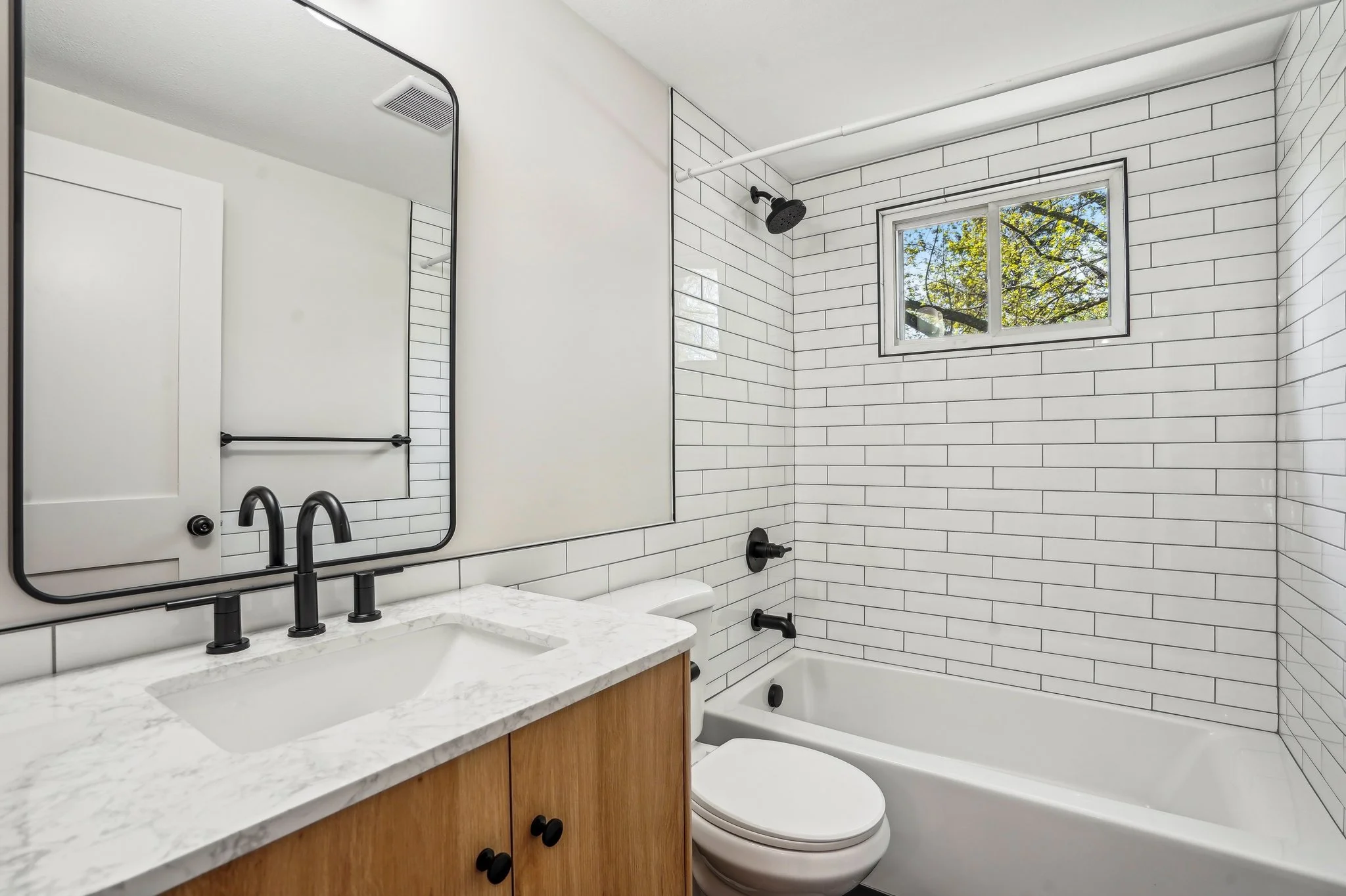 A modern bathroom with a white vanity, black faucet, large mirror, toilet, bathtub, and shower head. White subway tile walls and a small window showing trees outside.