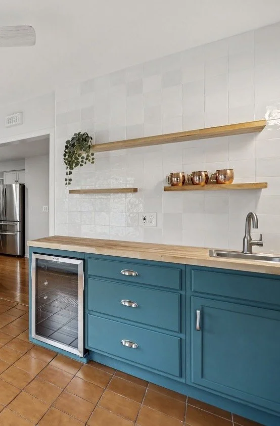 Kitchen with blue cabinets, wooden countertops, white tiled wall, open wooden shelves with copper cups and a hanging plant, and a wine cooler