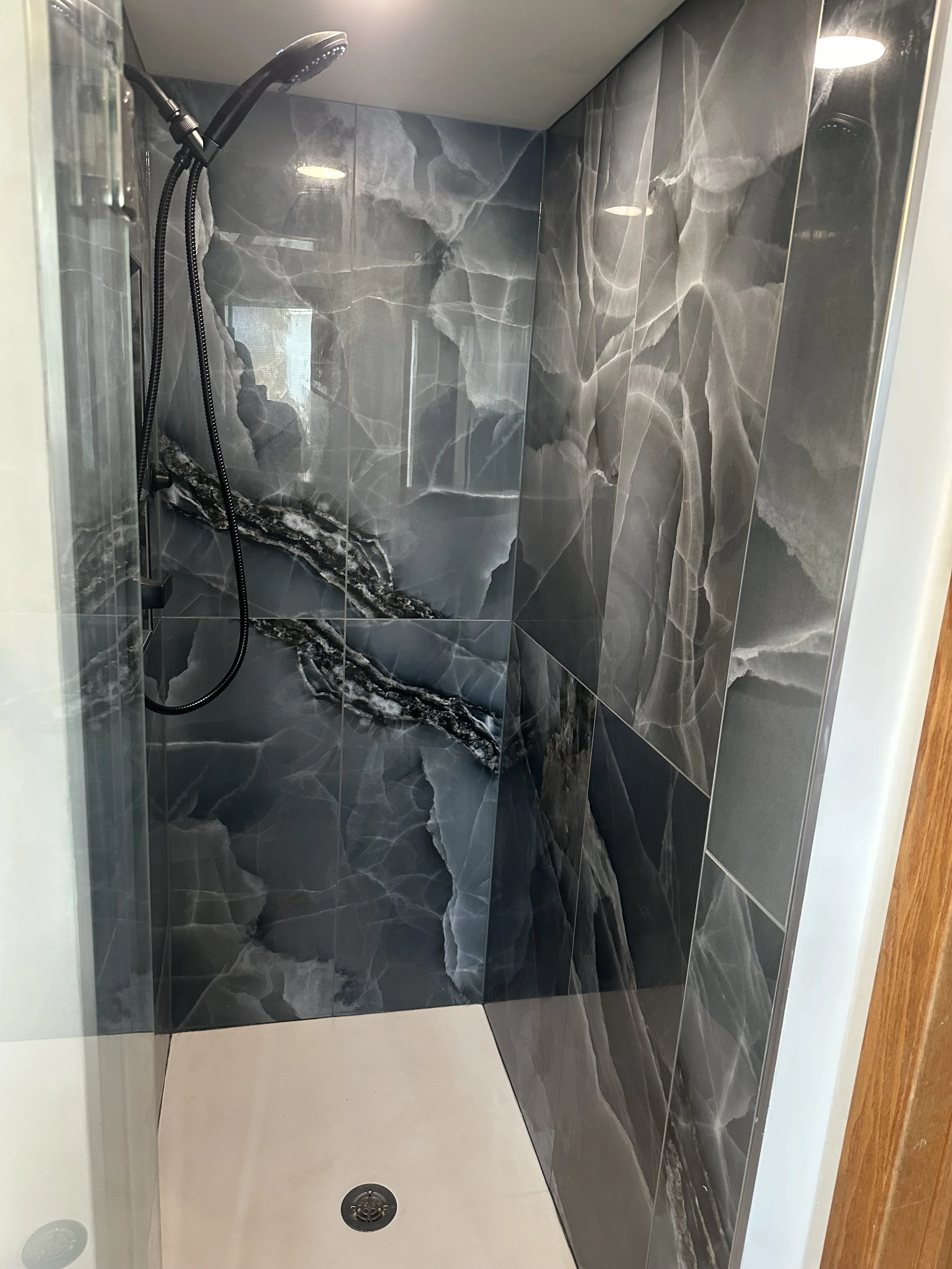 A walk-in shower with black and gray marble tiles, a handheld showerhead, and a drain in the beige floor.