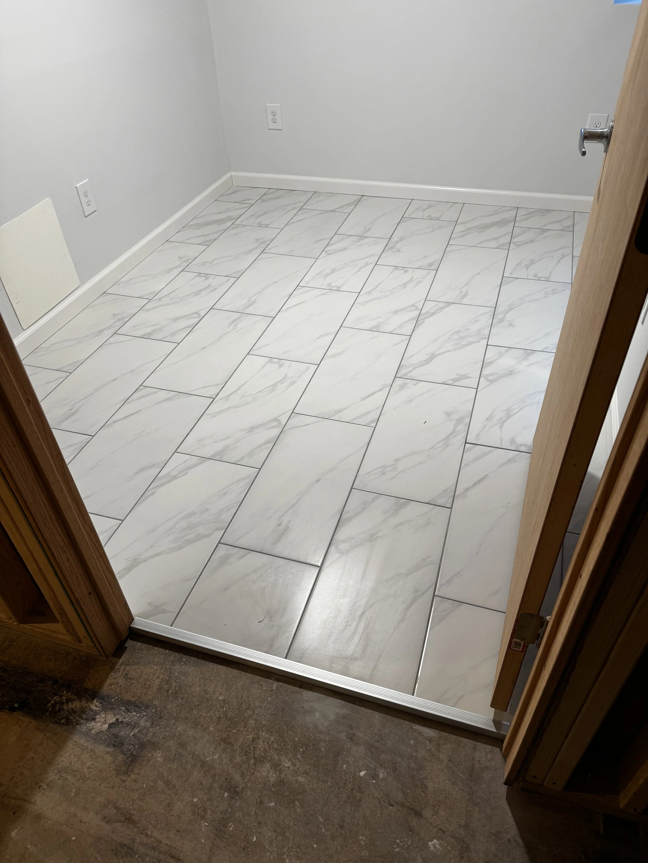 A small, empty room with white marble floor tiles, gray walls, a white baseboard, a wall outlet, a vent, and a partially open wooden door.