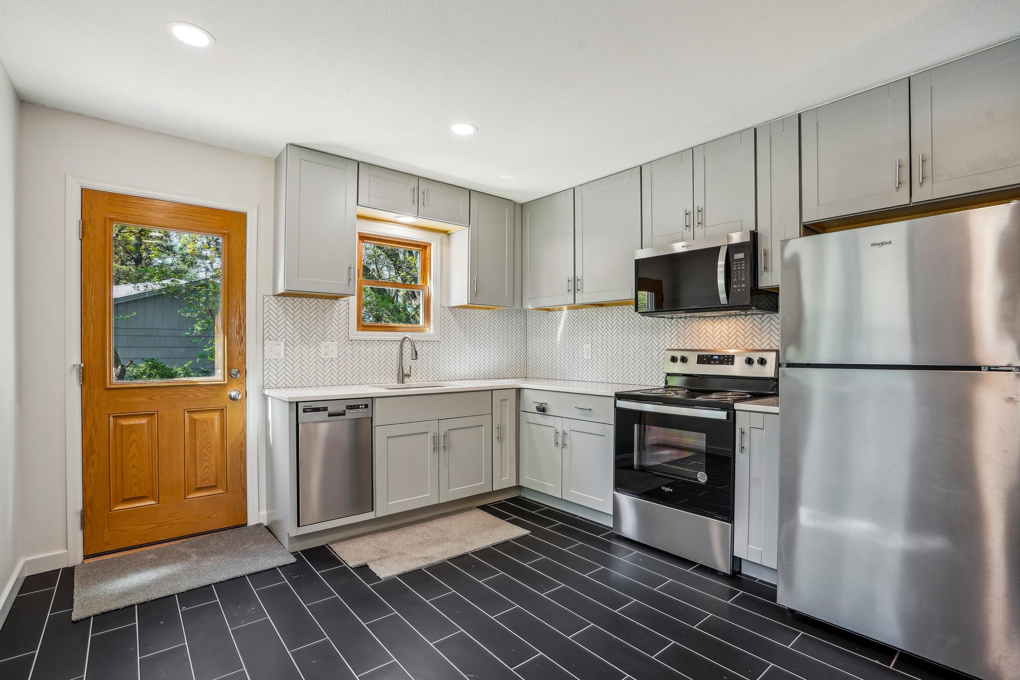 Kitchen with white cabinets, stainless steel refrigerator, stove, microwave, dishwasher, wooden door, two windows, black tile floor, and a neutral-colored backsplash.