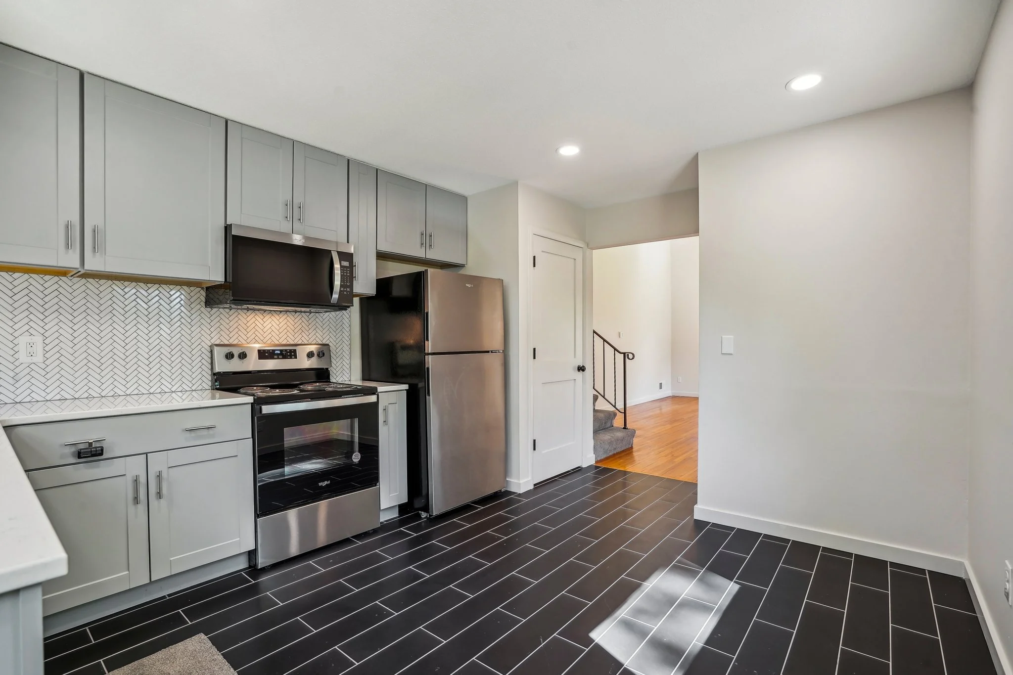 Modern kitchen with black tile flooring, light gray cabinets, stainless steel appliances including a refrigerator, oven, and microwave, white tile backsplash, and a view into a hallway with wood flooring and staircase.