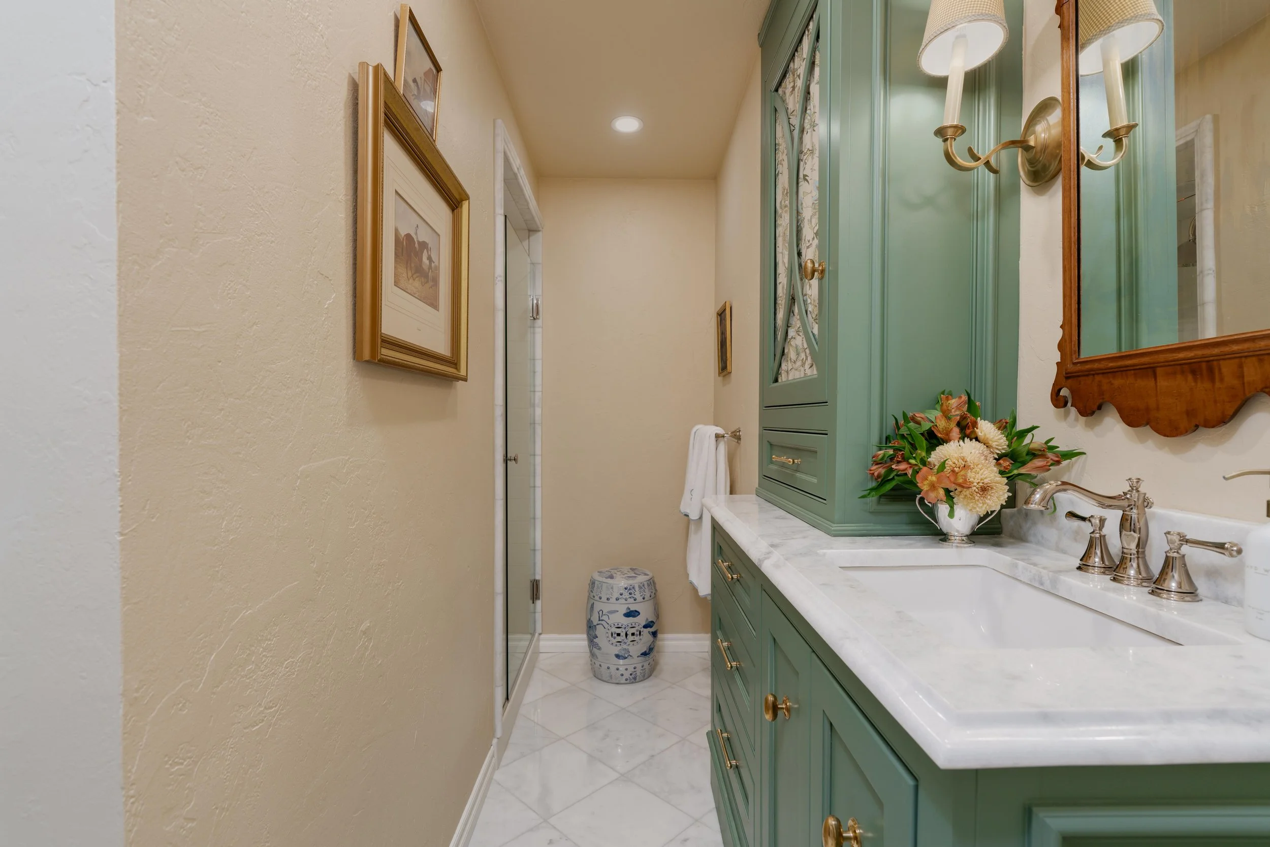 A bathroom with a green vanity and marble countertop, a white sink, silver fixtures, a flower arrangement, a mirror, wall-mounted light fixtures, framed artwork on textured beige walls, and a ceramic stool at the end of the room.