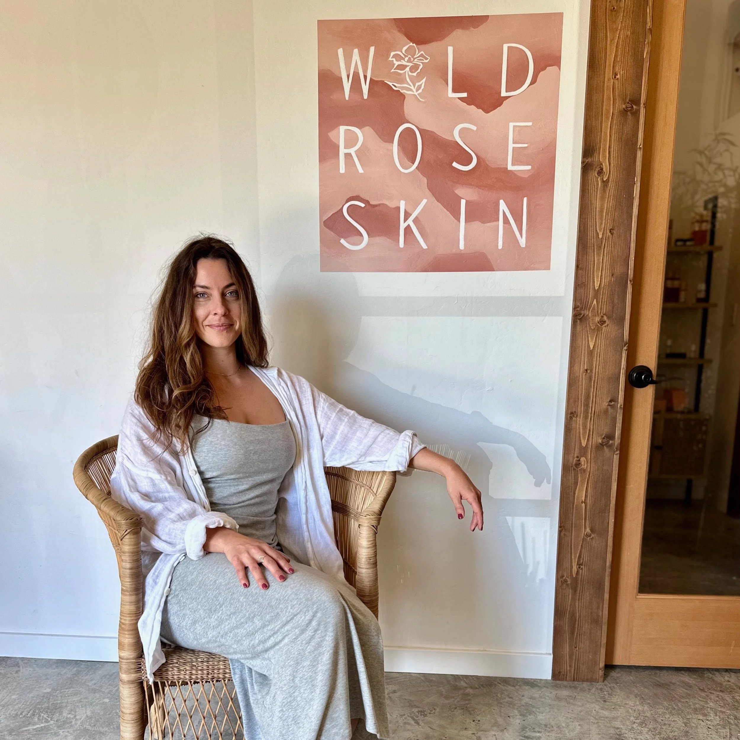A woman with long brown hair wearing a gray dress and white cover-up sitting on a wicker chair in front of a wall with a poster that reads 'Wild Rose Skin' in white letters on a pink background.