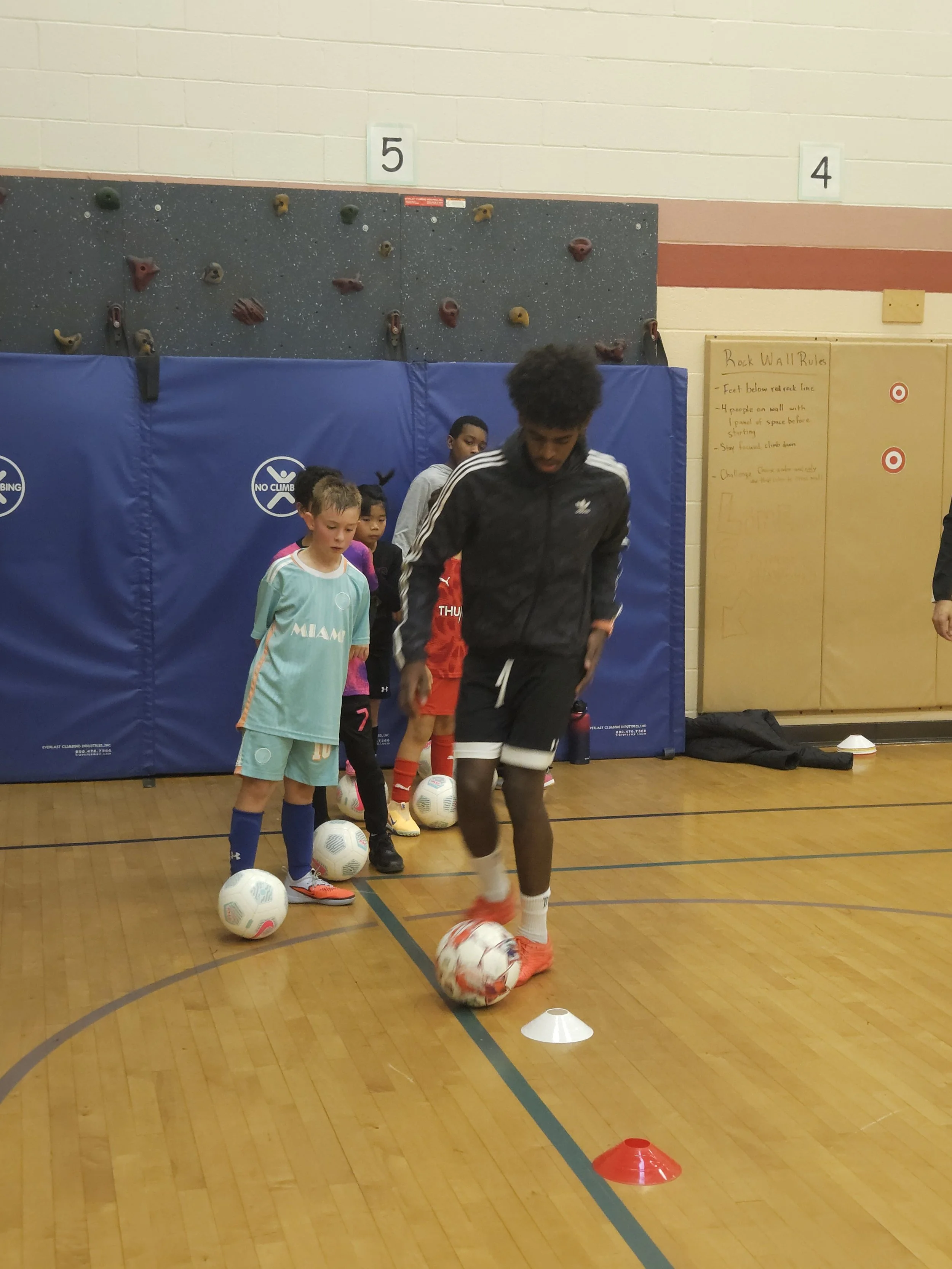 Children participating in indoor soccer training, with one boy dribbling a soccer ball through training cones while others wait in line.