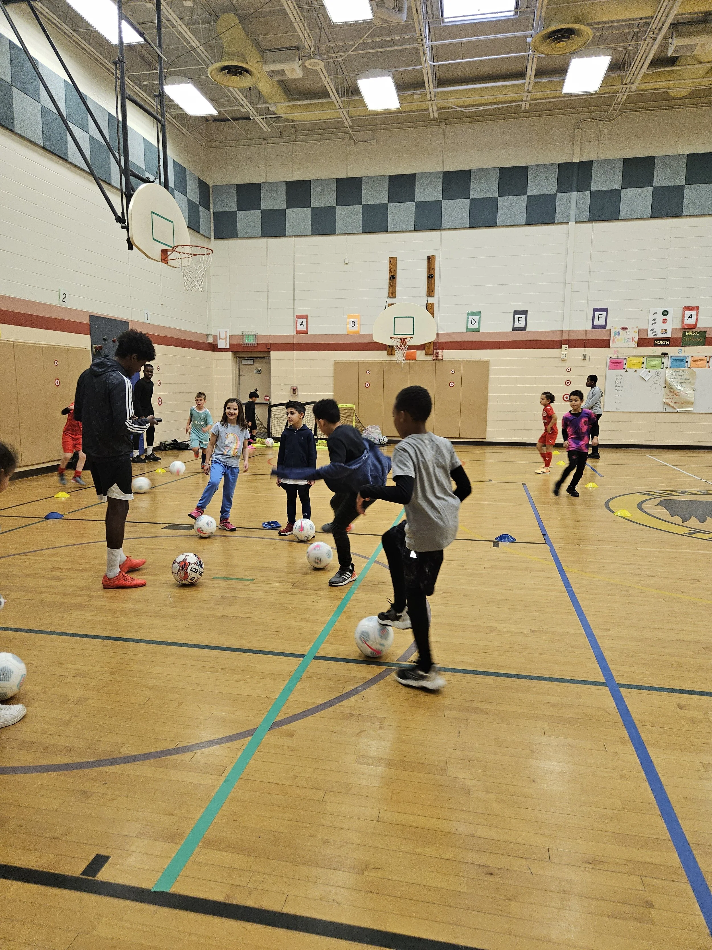 Children practicing soccer drills in a gymnasium with basketball hoops and colorful letters on the wall.