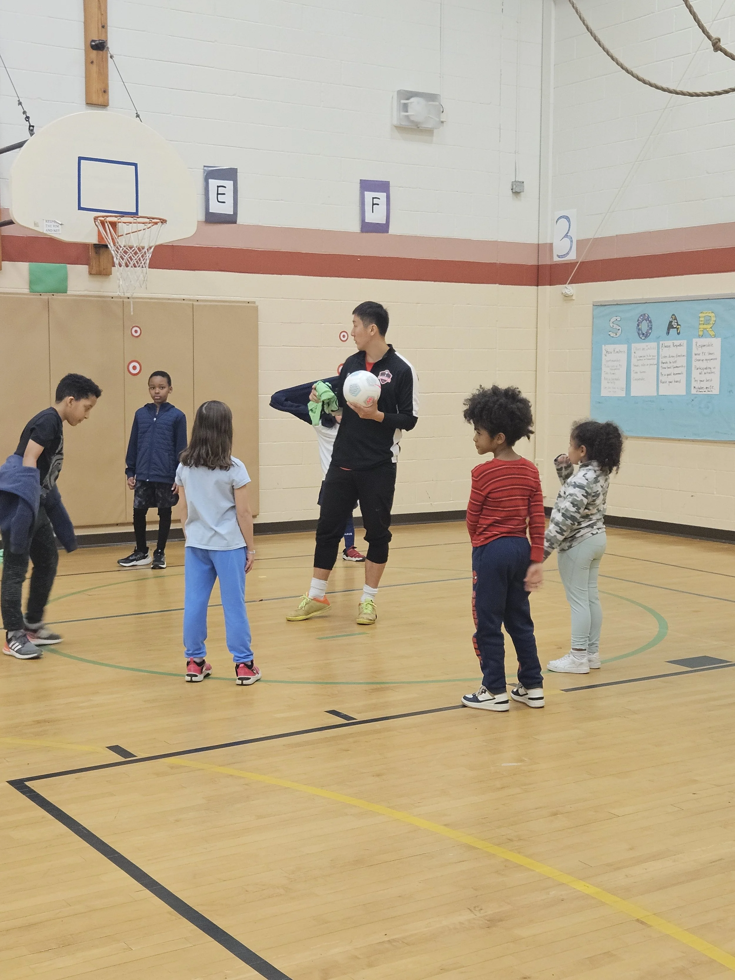 A group of children gathered around an adult coach in a gymnasium, seemingly preparing for or participating in a basketball practice or game. The coach is holding a basketball and some other items.