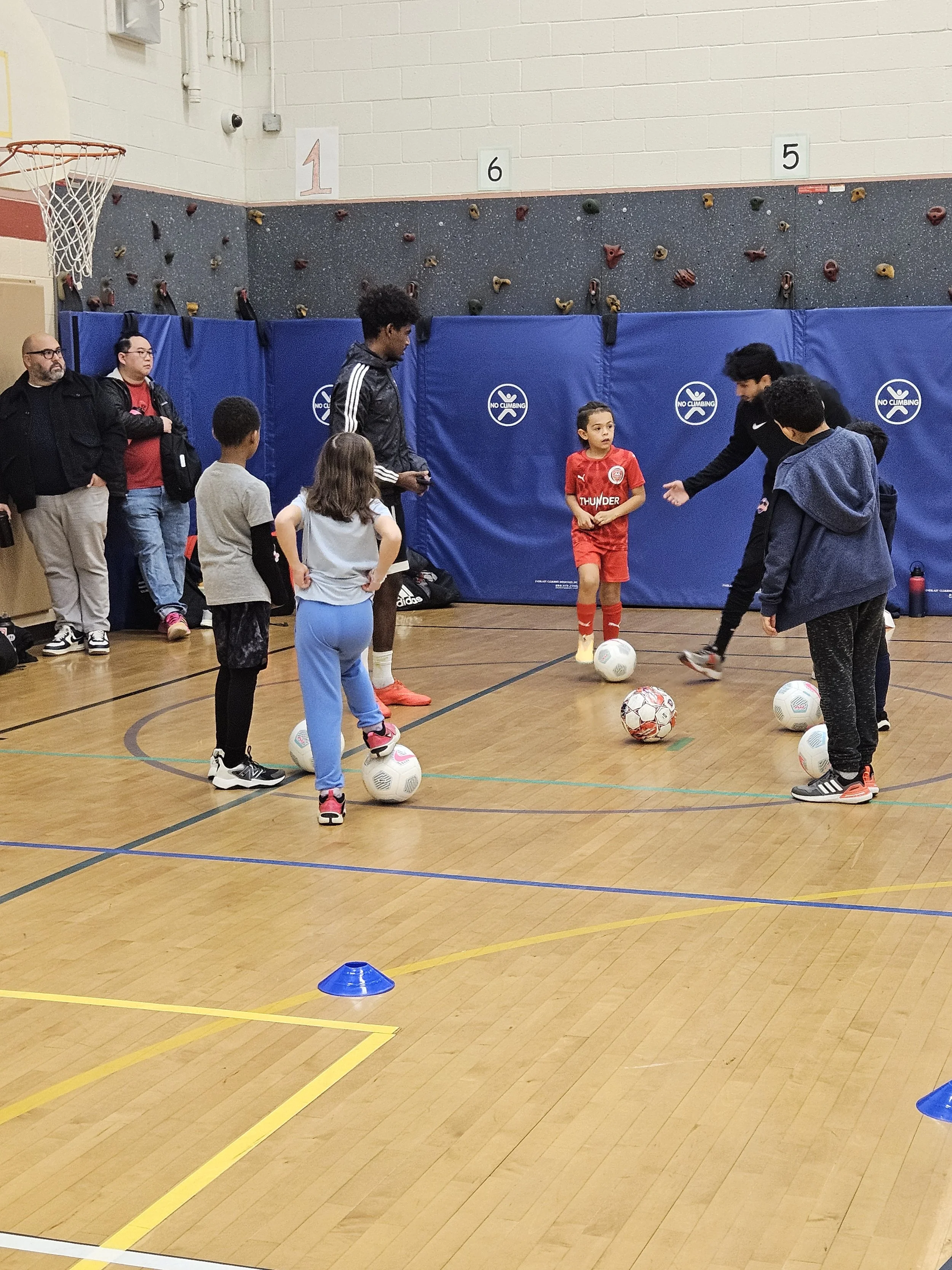 Children participating in a soccer drill in an indoor gym, with coaches and adult observers present, and rock climbing wall in the background.