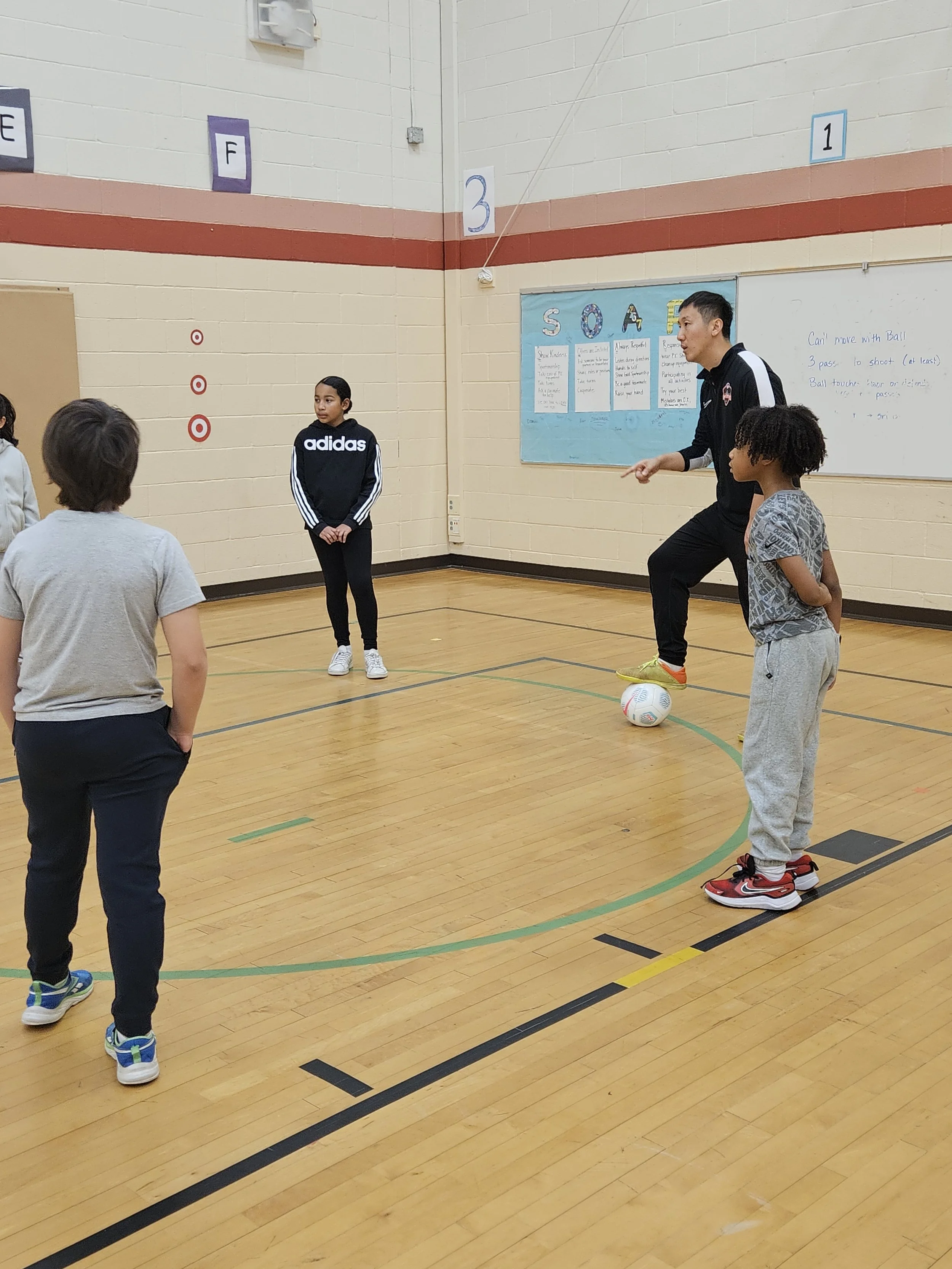 Children participating in an indoor soccer class with an instructor, who is demonstrating a soccer drill. The instructor is balancing on a soccer ball, giving instructions to the group in a gymnasium.