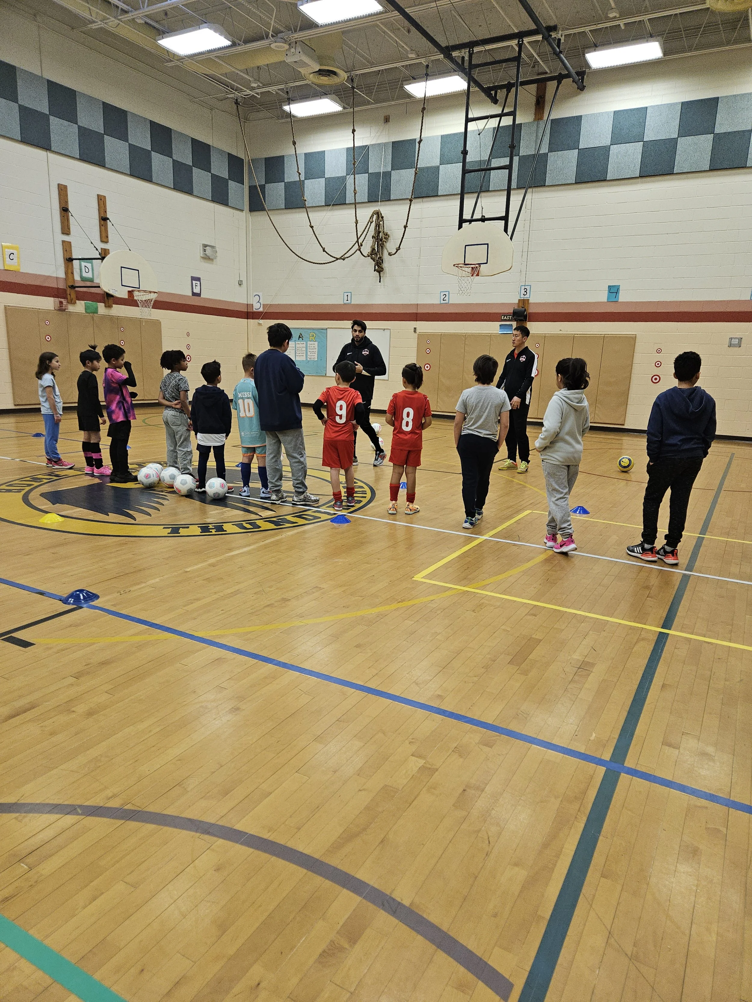 Children are gathered in a gymnasium for a sports lesson, with two coaches giving instructions. Some children are in athletic clothing, some are in casual clothes, and soccer balls are on the floor. The gym has basketball hoops, climbing ropes, and a