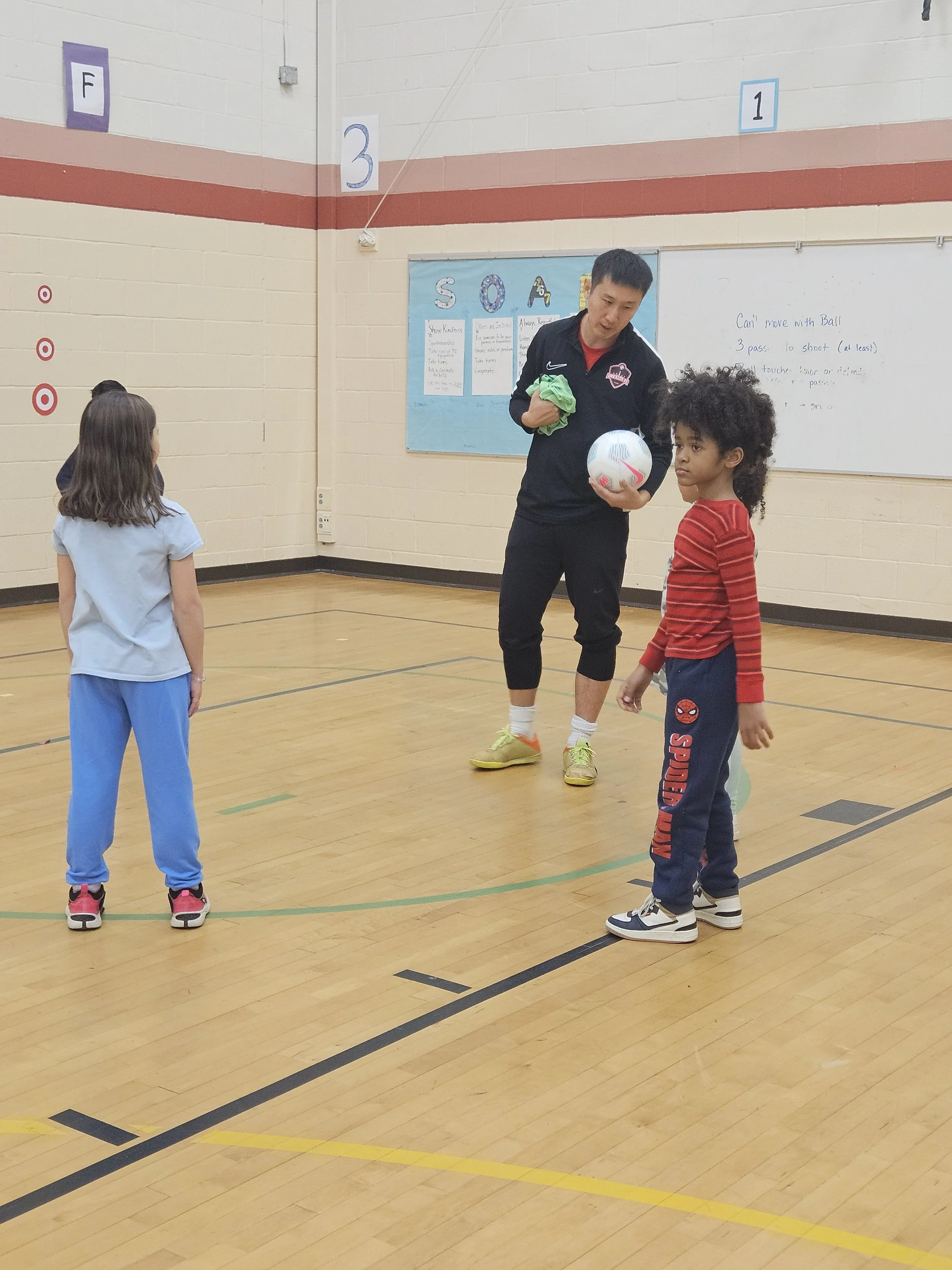 A coach demonstrating soccer skills with two children in a gymnasium, holding a soccer ball and a green cloth, with a whiteboard and bulletin board in the background.
