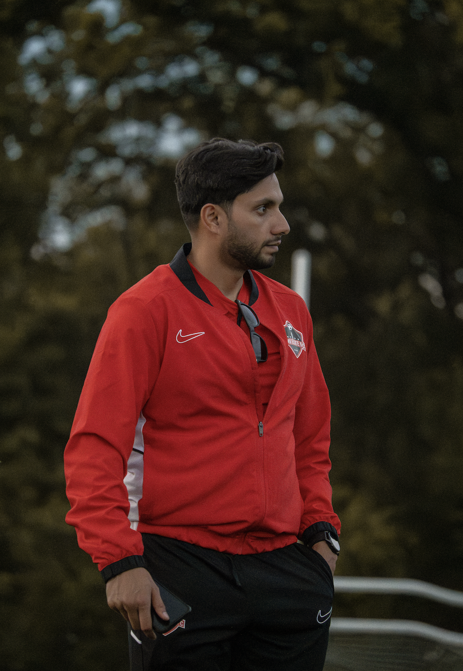 A man in a red shirt holding a tactical football strategy board with diagrams and markers, standing in front of a large football club logo with a soccer ball symbol.