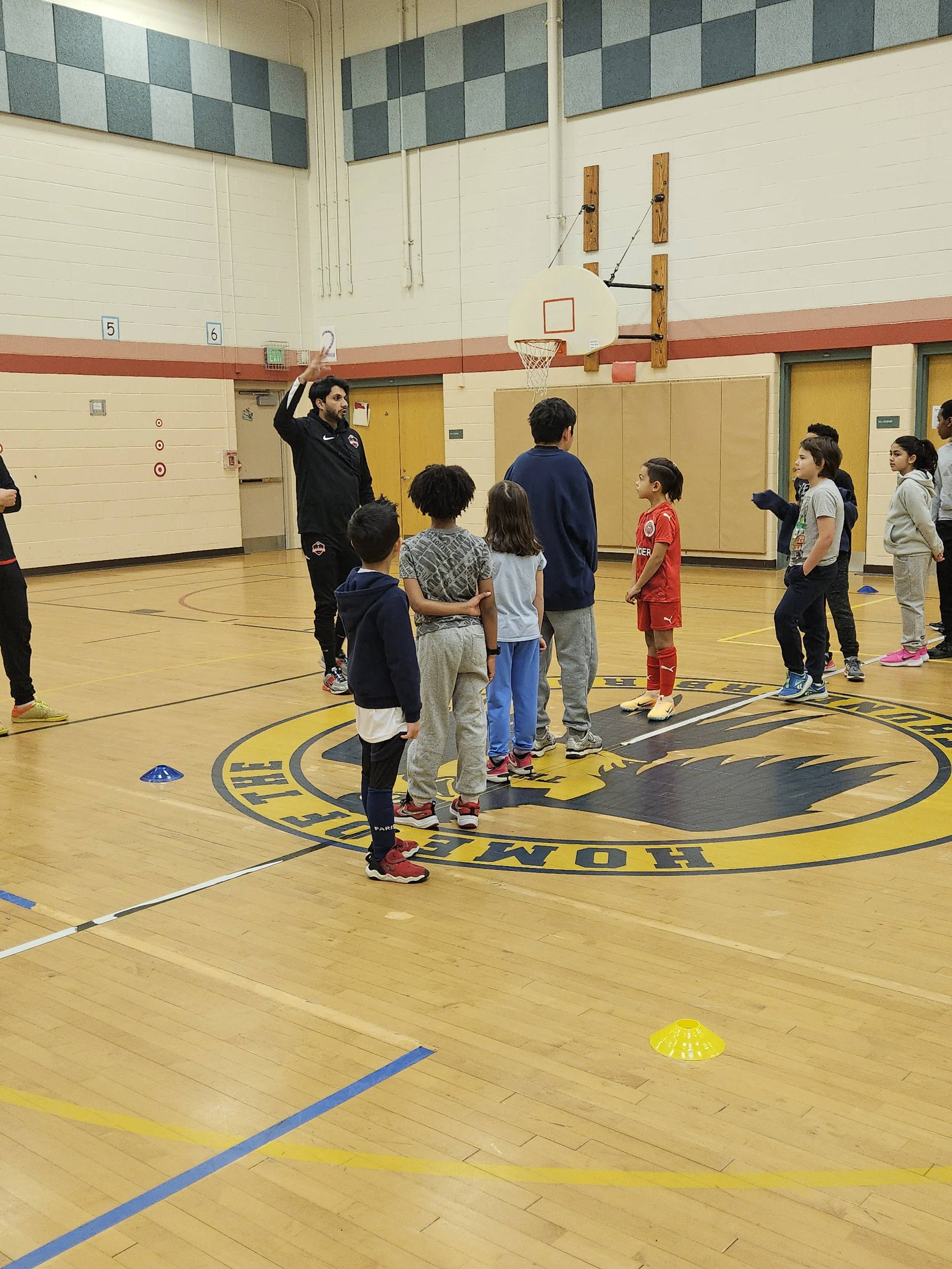 A basketball coach giving instructions to a group of young children on a basketball court, with children lined up and listening.