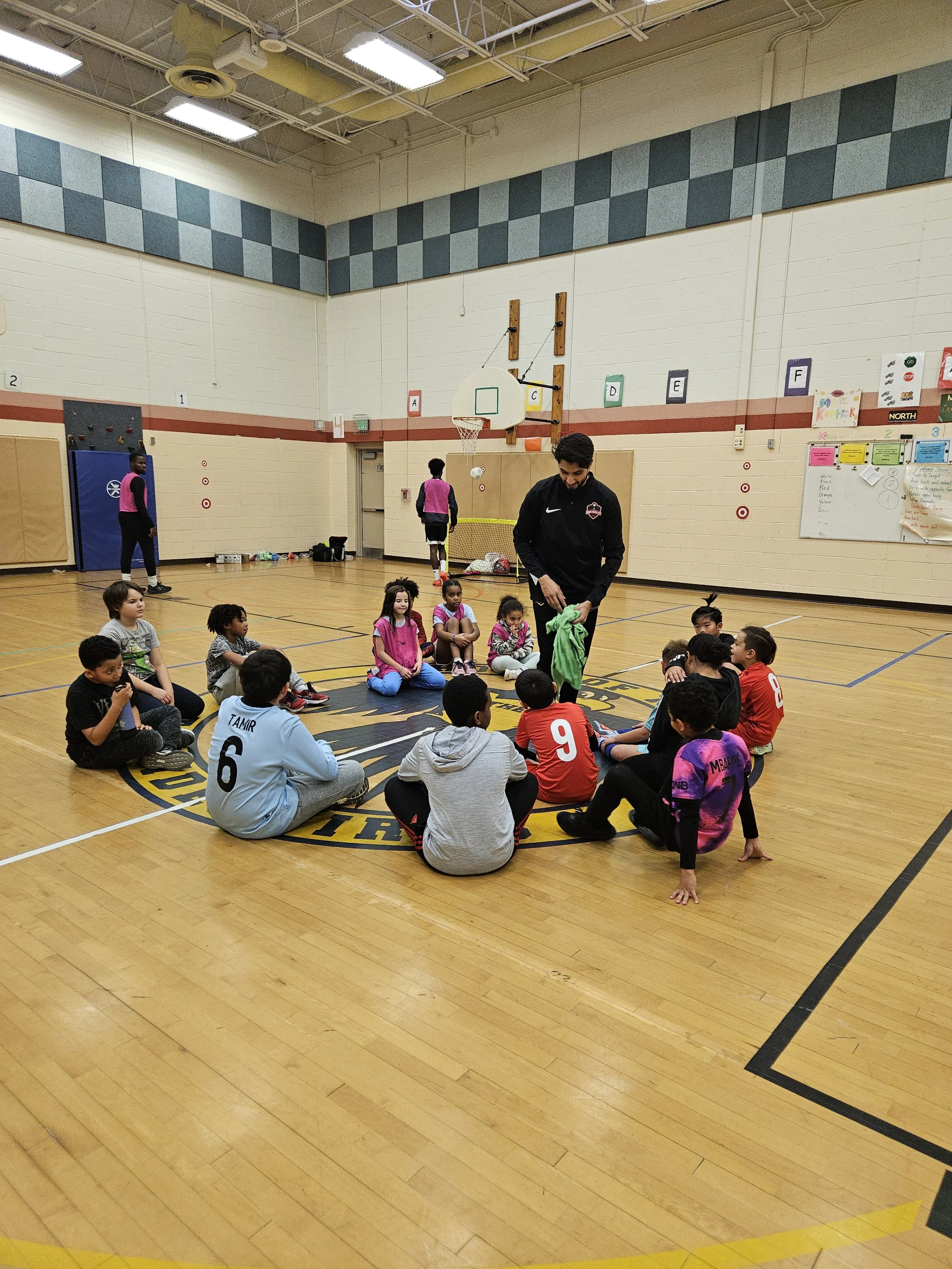 A coach standing in the center of a circle of young children sitting on a basketball court in a gymnasium. The children are wearing sports jerseys and casual clothing. Some children are sitting on the floor while others are standing or kneeling nearb