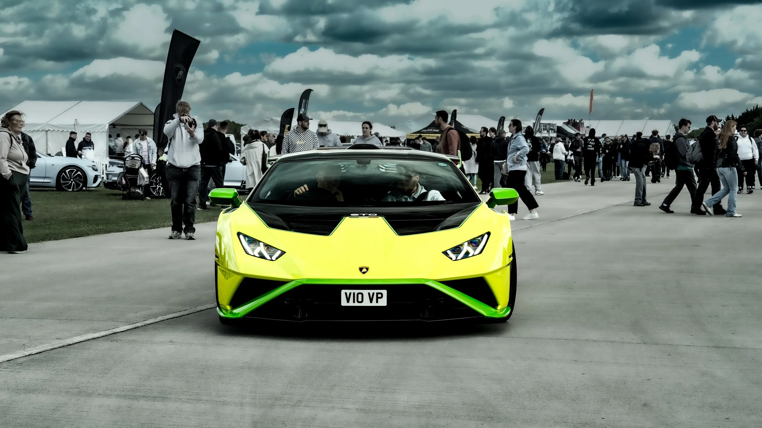 A bright yellow and black Lamborghini sports car on display at an outdoor event with many people walking around and tents in the background under a cloudy sky.