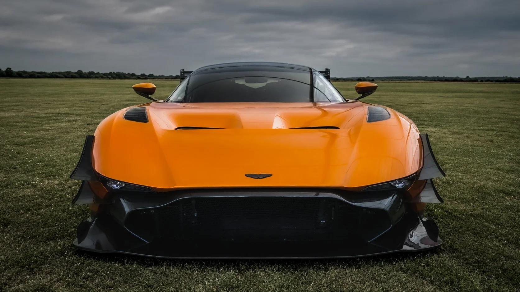 Front view of an orange high-performance sports car on a grassy field with cloudy sky.