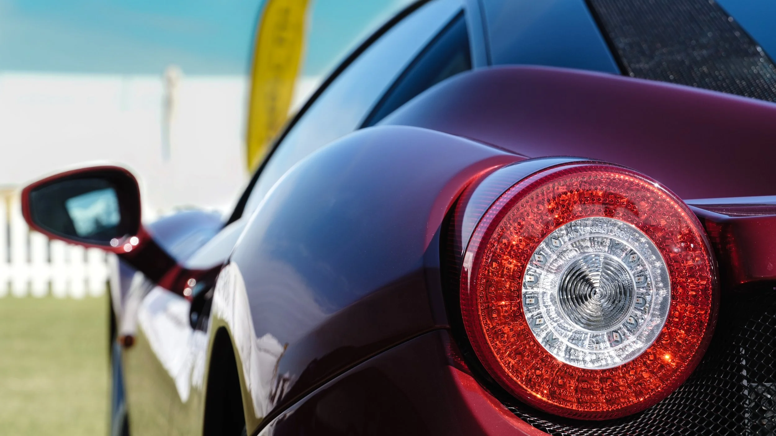 Close-up of the rear end of a sleek black sports car, showing its round tail light with LED detailing and part of the side mirror, parked outdoors on a sunny day.