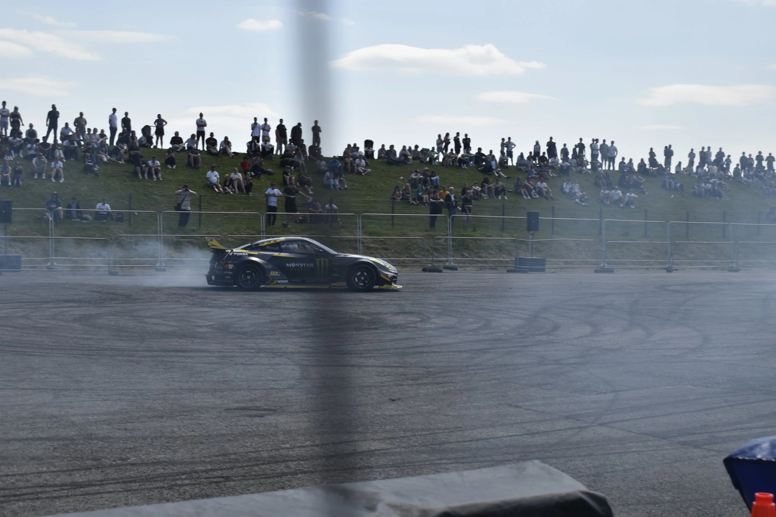 A race car speeding on a track with a crowd of spectators sitting and standing on a grassy hill in the background.