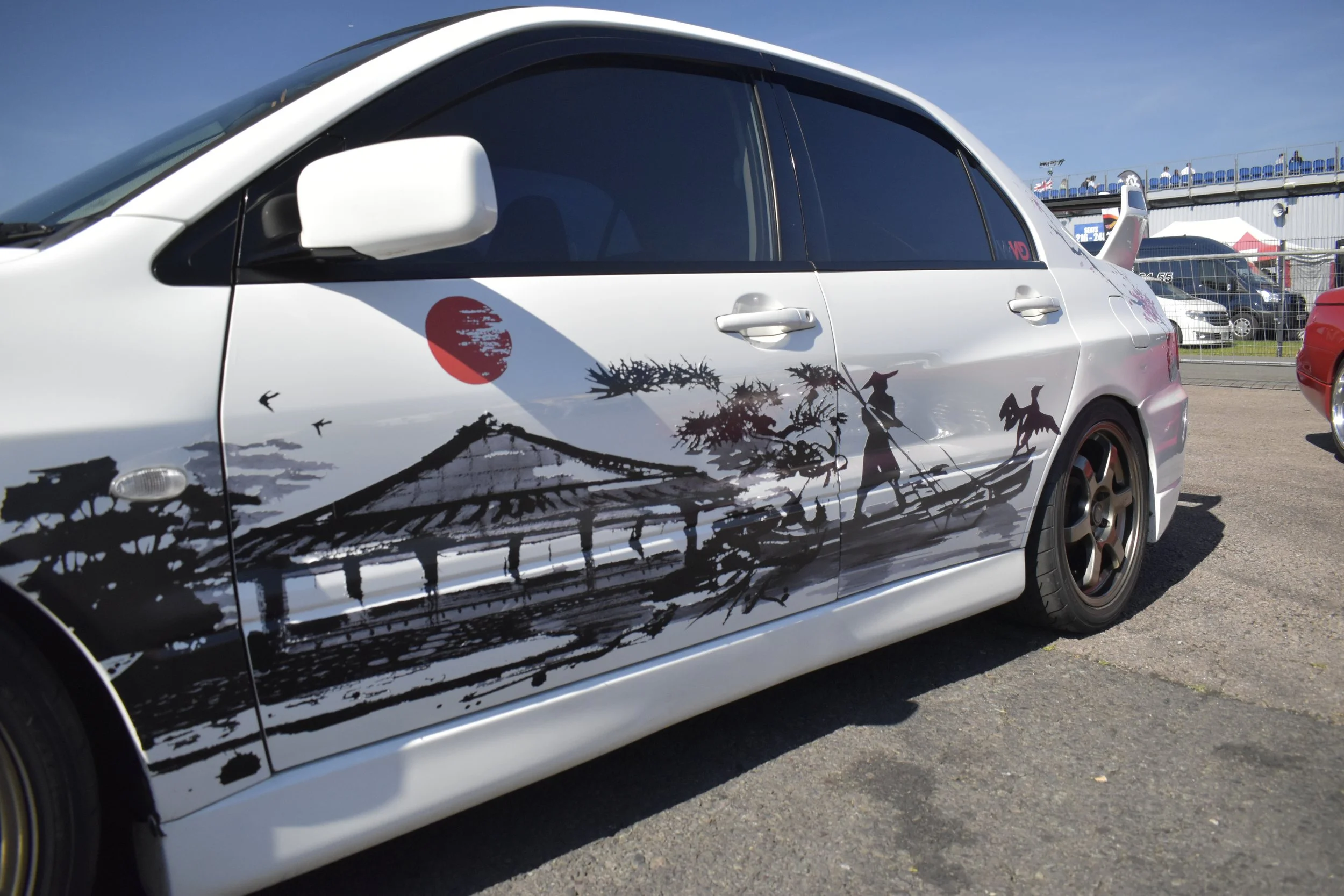 A white car with a black and white Japanese-themed graphic featuring a red sun, trees, and birds on its side panel, parked outdoors on a sunny day.