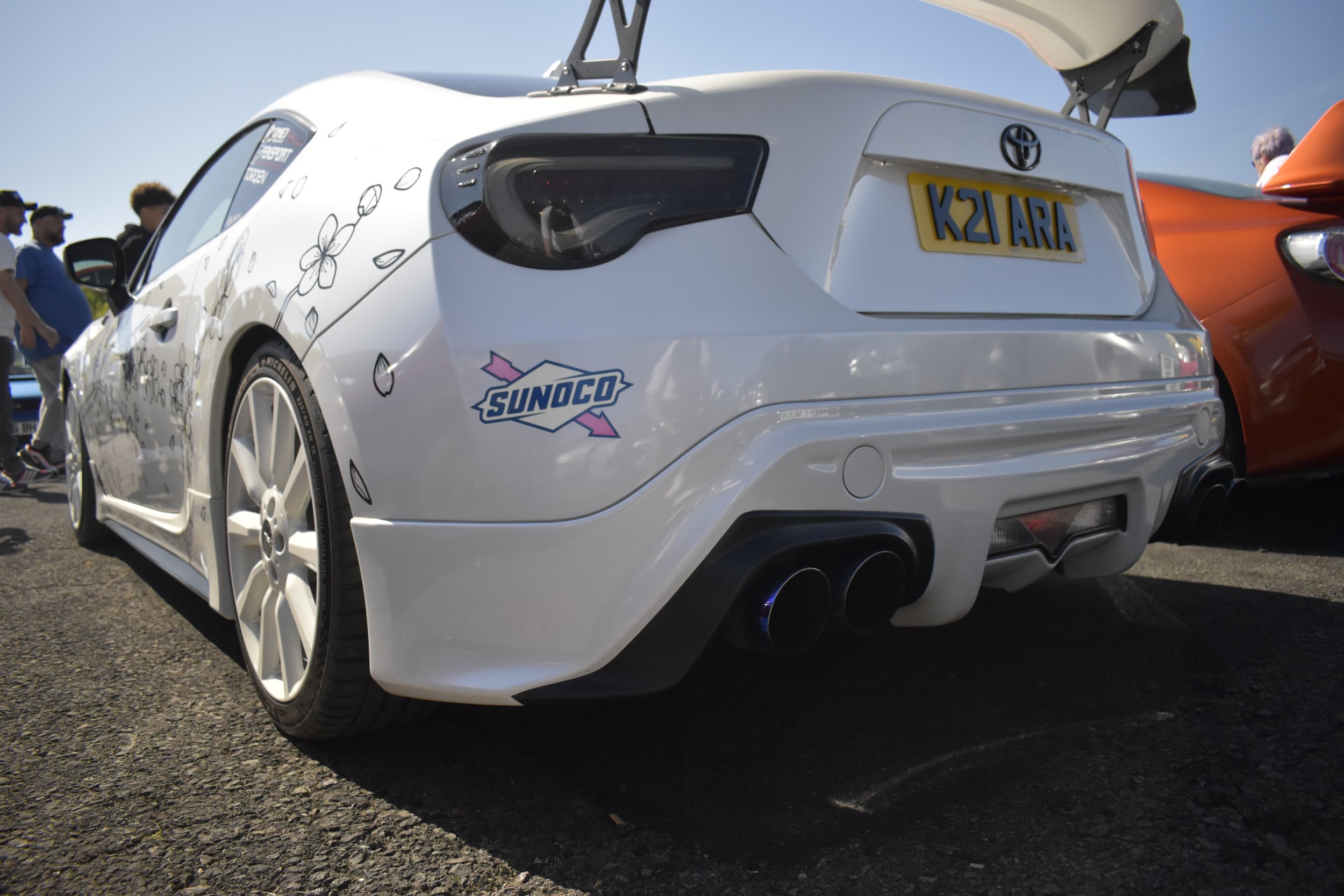 Rear view of a white sports car with custom decals, a large rear wing, and a Japanese license plate reading 'K21 ARA', parked at an outdoor event with a group of people in the background.