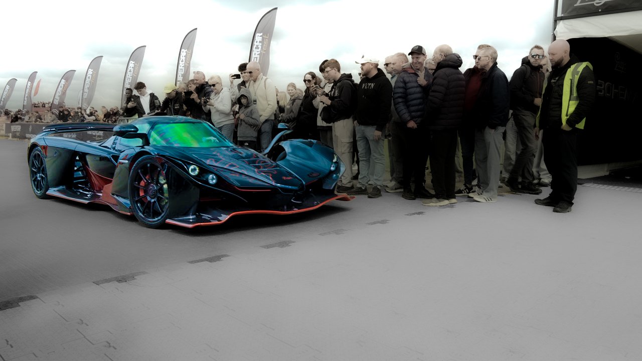 A line of spectators watching a futuristic black and red sports car on display at an outdoor event with cloudy skies.