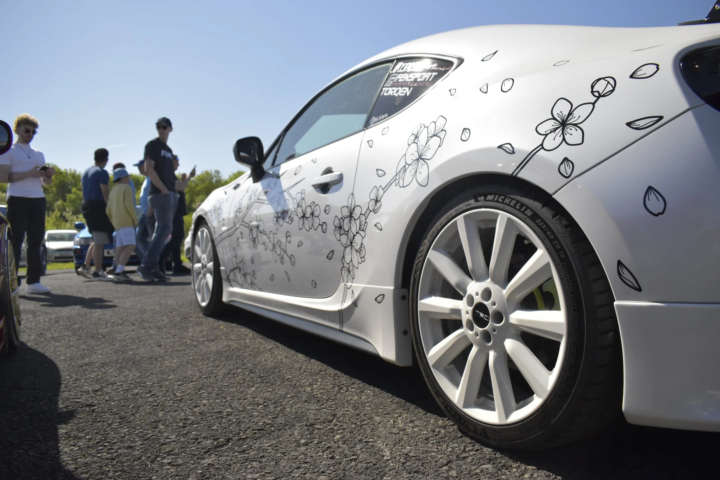 Close-up of a white sports car with floral decals parked outdoors, with a group of people in the background and a clear blue sky.