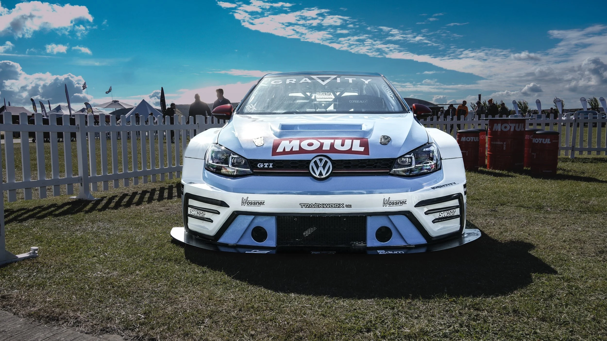 A white Volkswagen GTI race car with Motul sponsorship, parked on a grass field at a motorsport event, with tents and people in the background under a partly cloudy sky.