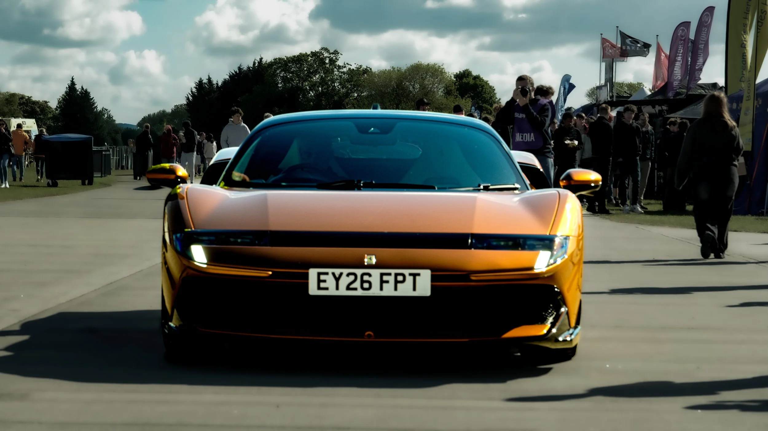 A gold-colored sports car driving on a runway or road with a crowd of people and tents in the background.