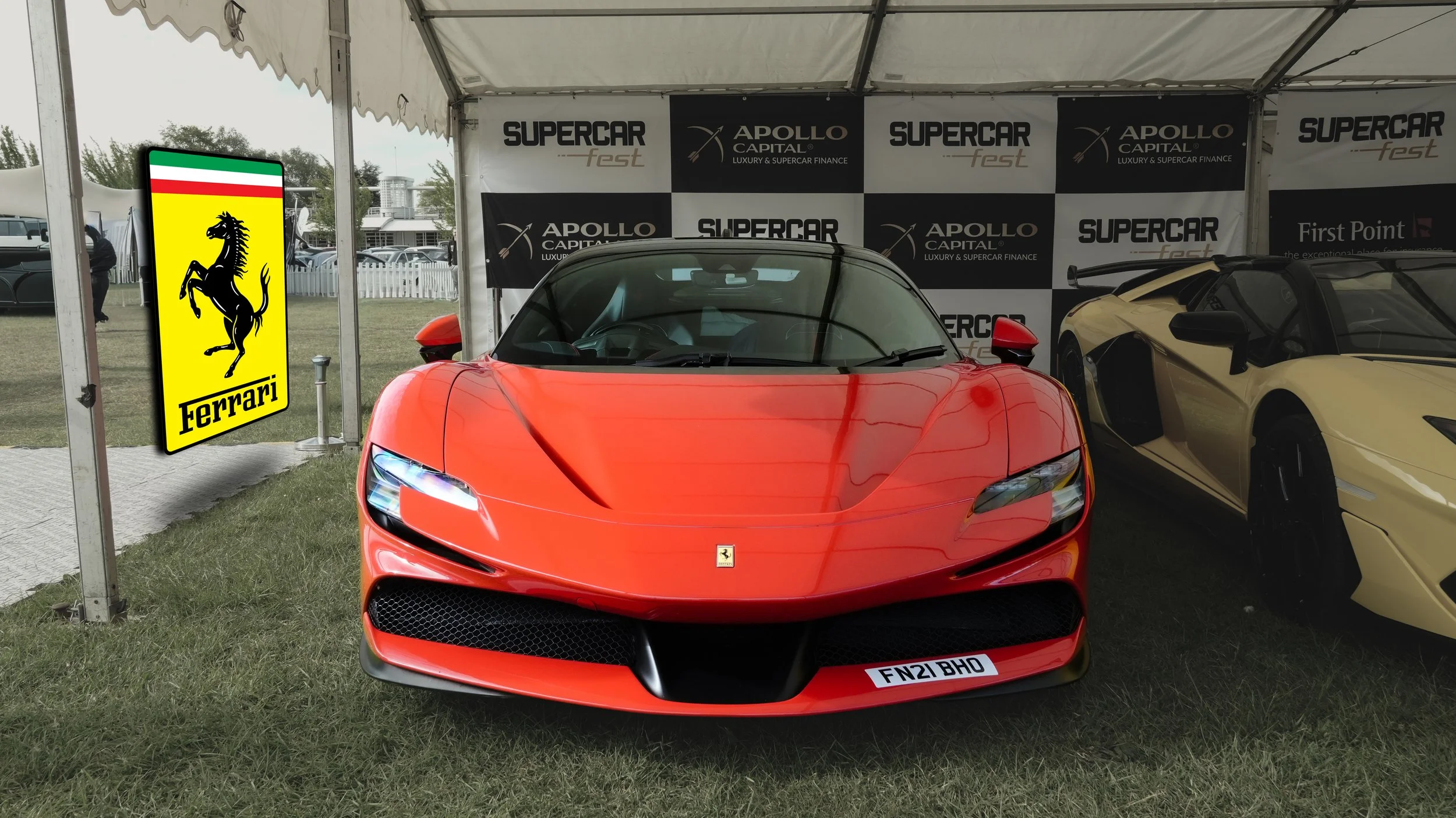 Red Ferrari sports car with black accents displayed under a tent at a car event, with a Ferrari logo sign to the left and a black and white checkered backdrop behind.