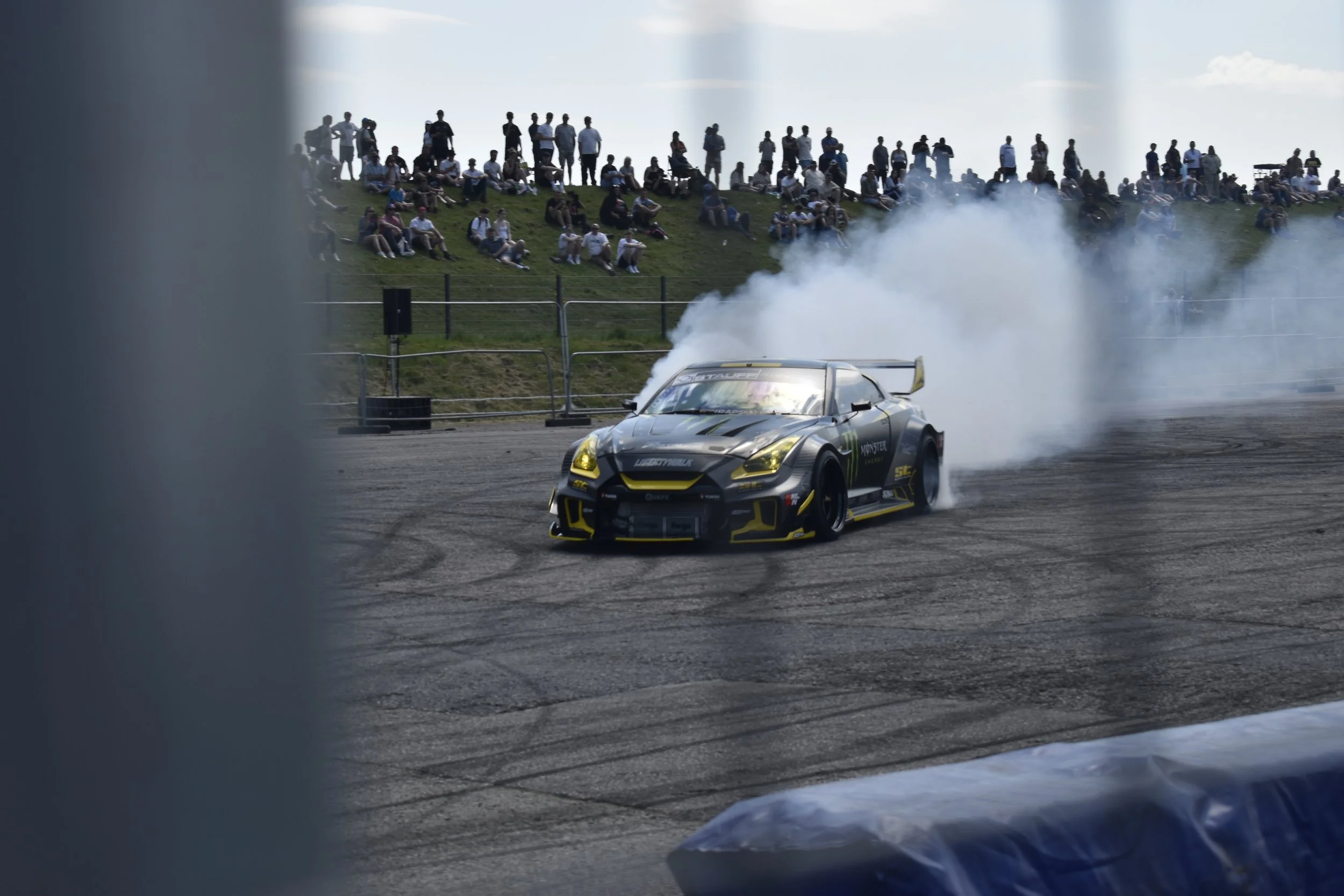 A race car with Monster Energy branding drifting on a track, with smoke trailing behind it, viewed through a partially obstructed perspective, and a crowd of spectators sitting and standing on a grassy hillside in the background.