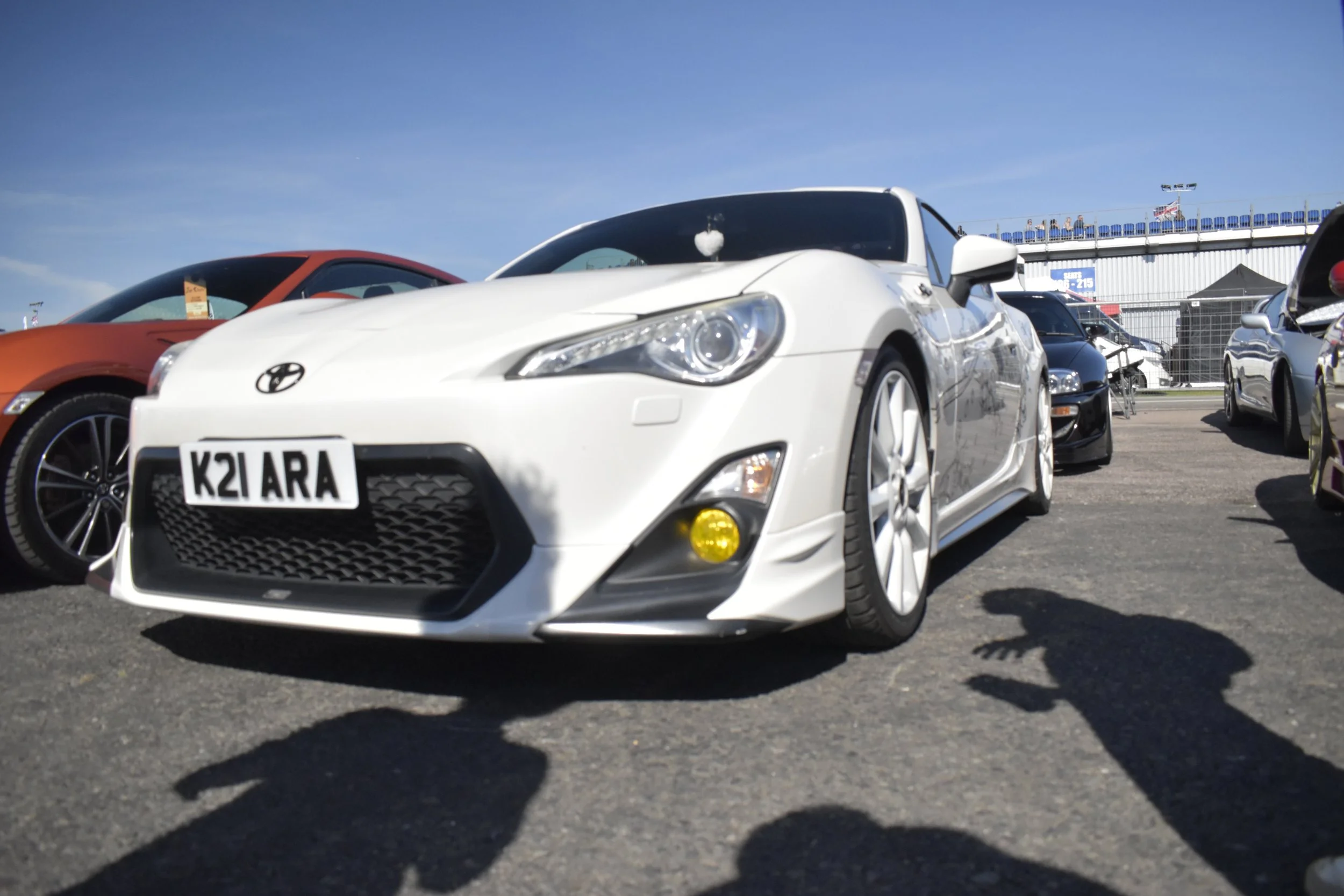 A white Toyota sports car with a custom license plate K21 ARA parked at a car show, with other cars and a fence in the background under a clear blue sky.