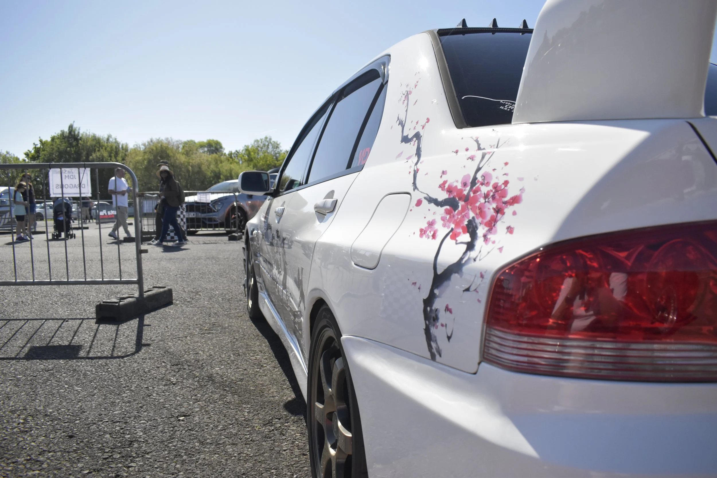 Close-up of a white car with pink cherry blossom design decals on the side, parked outdoors near a black metal fence, with people and other vehicles in the background under a clear blue sky.