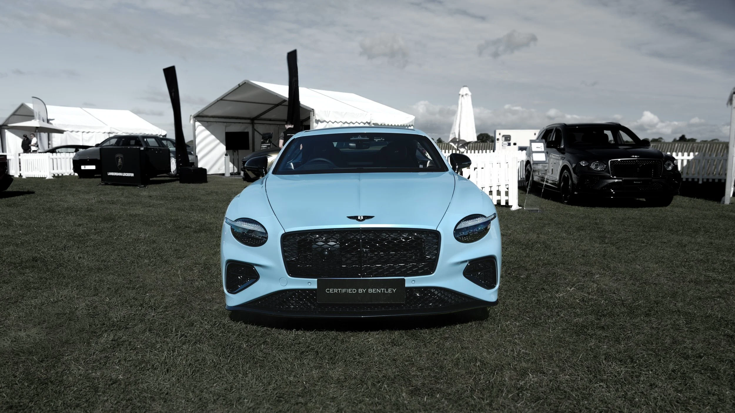 Light blue luxury car showcase at an outdoor event with white tents and black cars in the background.