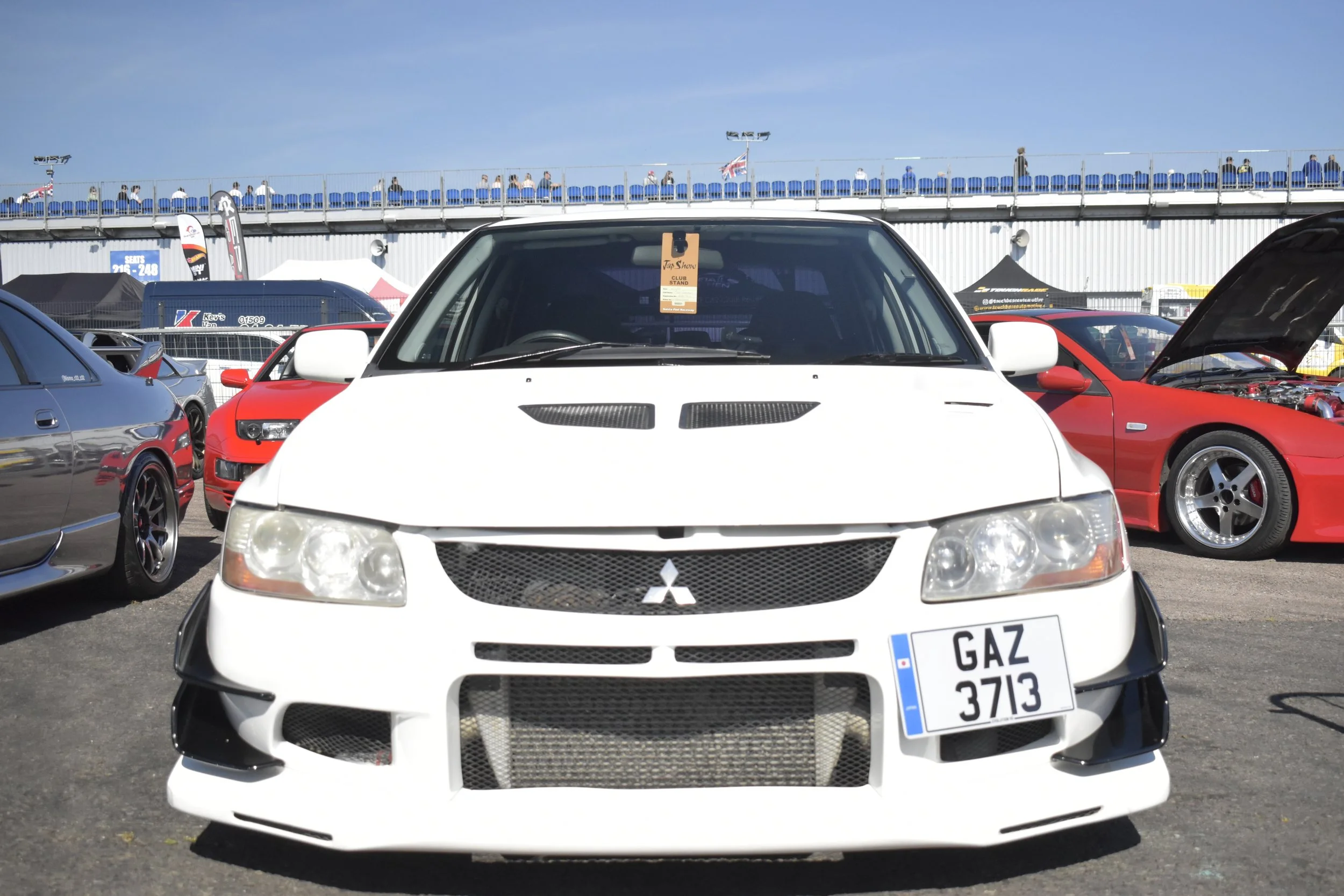 Front view of a white Mitsubishi Lancer Evo with a European license plate, parked among other cars at a racing event or car show, with tents and spectators visible in the background.