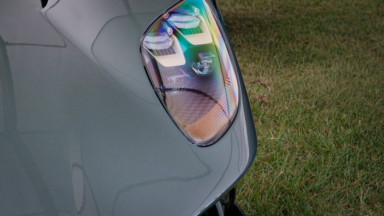 Close-up of a car headlight on a silver vehicle parked on grass, with visible reflections on the car's surface.