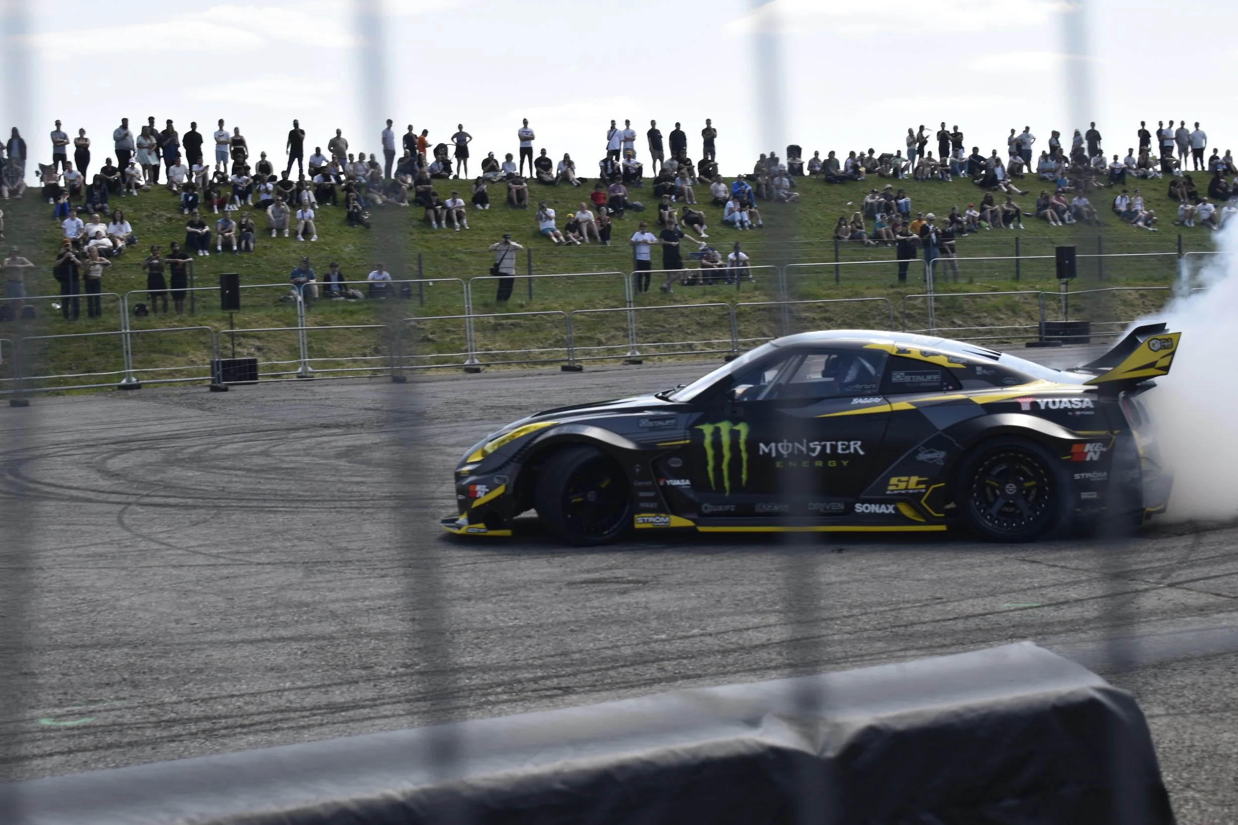 Race car with Monster Energy and other sponsor logos speeding on a track, while spectators watch from a grassy hillside.