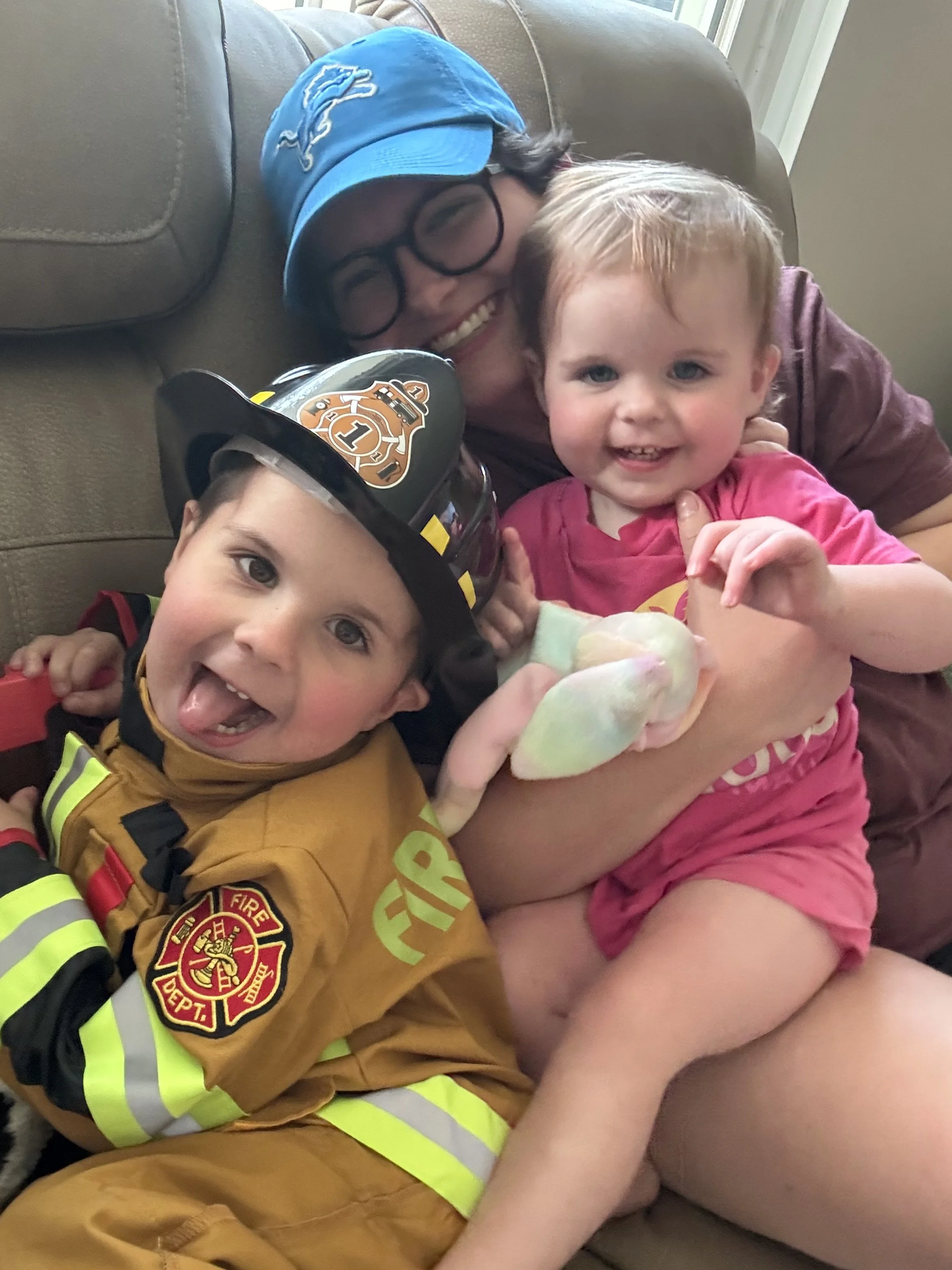 A firefighter in uniform and a girl are lying on a couch, smiling and hugging. The firefighter is wearing a yellow uniform with reflective stripes and a helmet. The girl has blonde hair, blue eyes, and is wearing a pink shirt and shorts.